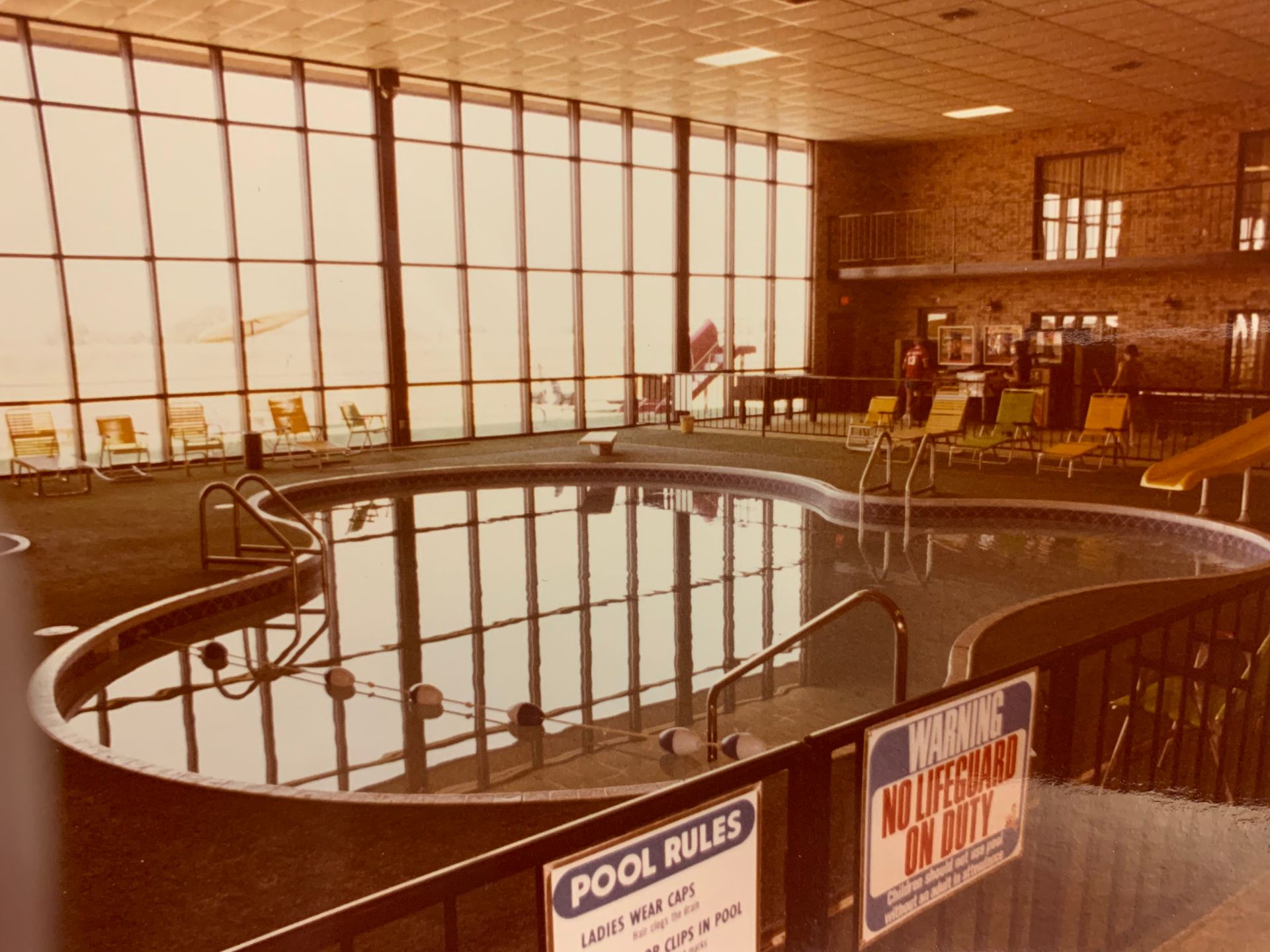 Indoor swimming pool with large windows, lounge chairs, and a small slide. Pool rules sign in foreground.