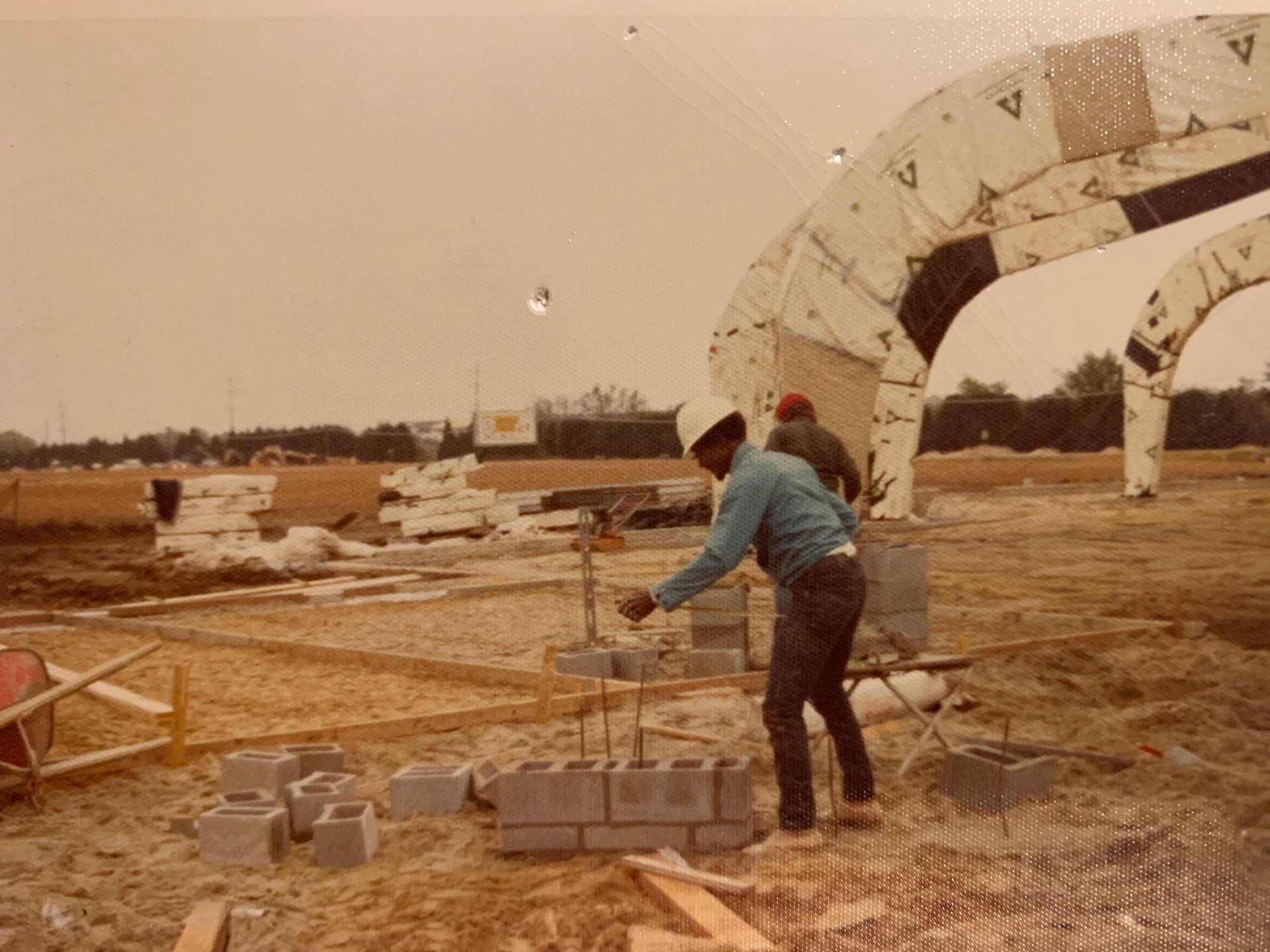 Construction worker builds a brick structure outdoors, near a large inflatable structure and a partially cleared lot.
