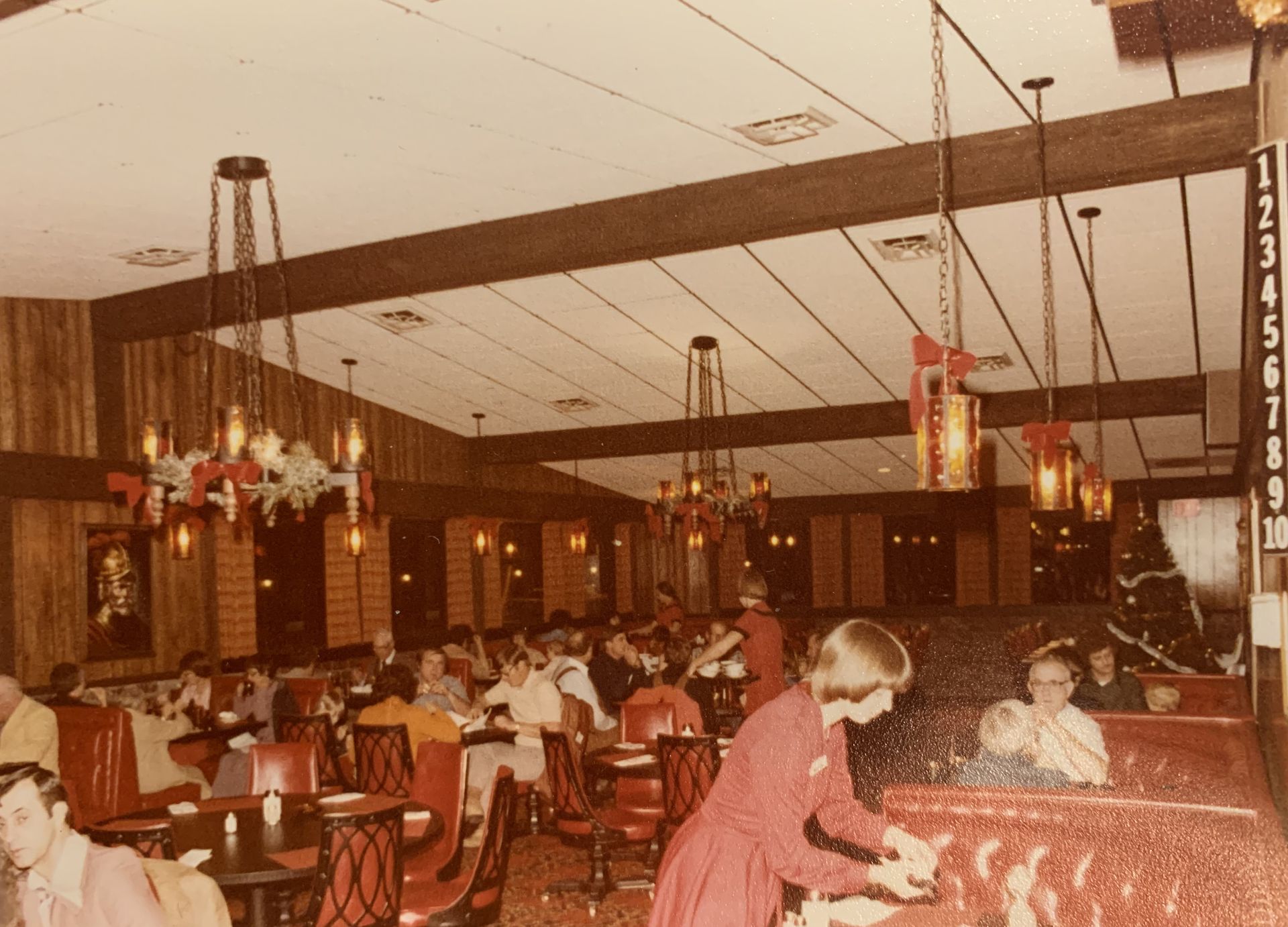 Interior view of a vintage restaurant with patrons seated at tables. Servers in red uniforms are visible.