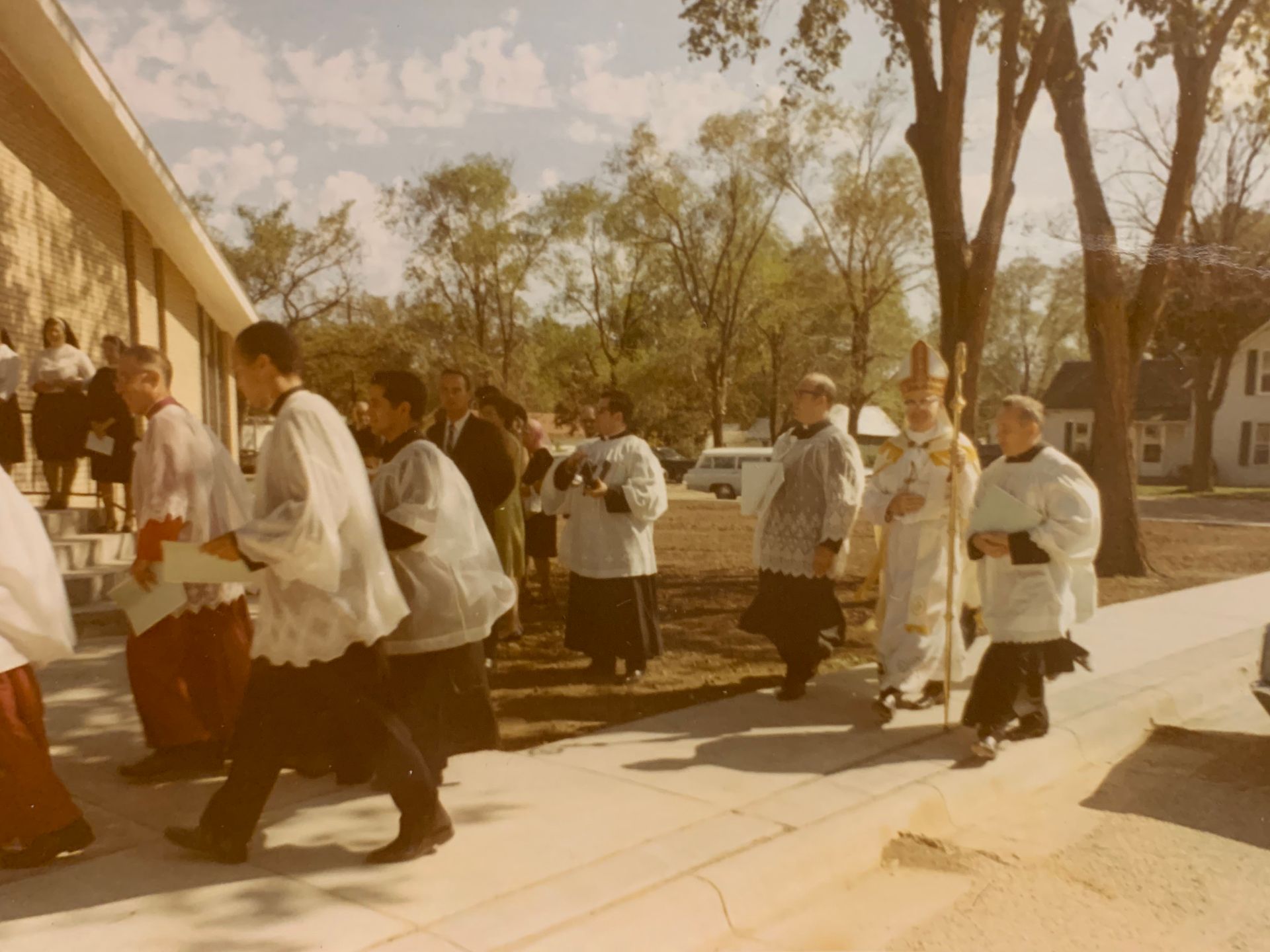 Clergy in procession outside a building; white robes, tall cross, trees in background, sunny day.