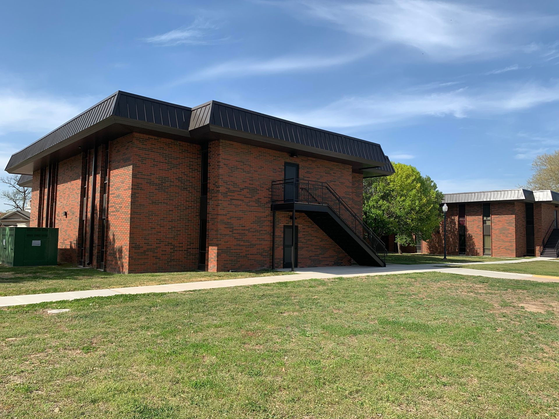 Two brick buildings with dark roofs and a grassy area under a blue sky.