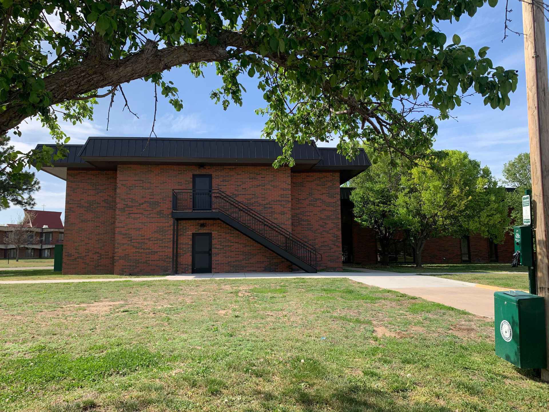 Brick building with a dark metal staircase, trees, and a green trash can on a grassy lawn.