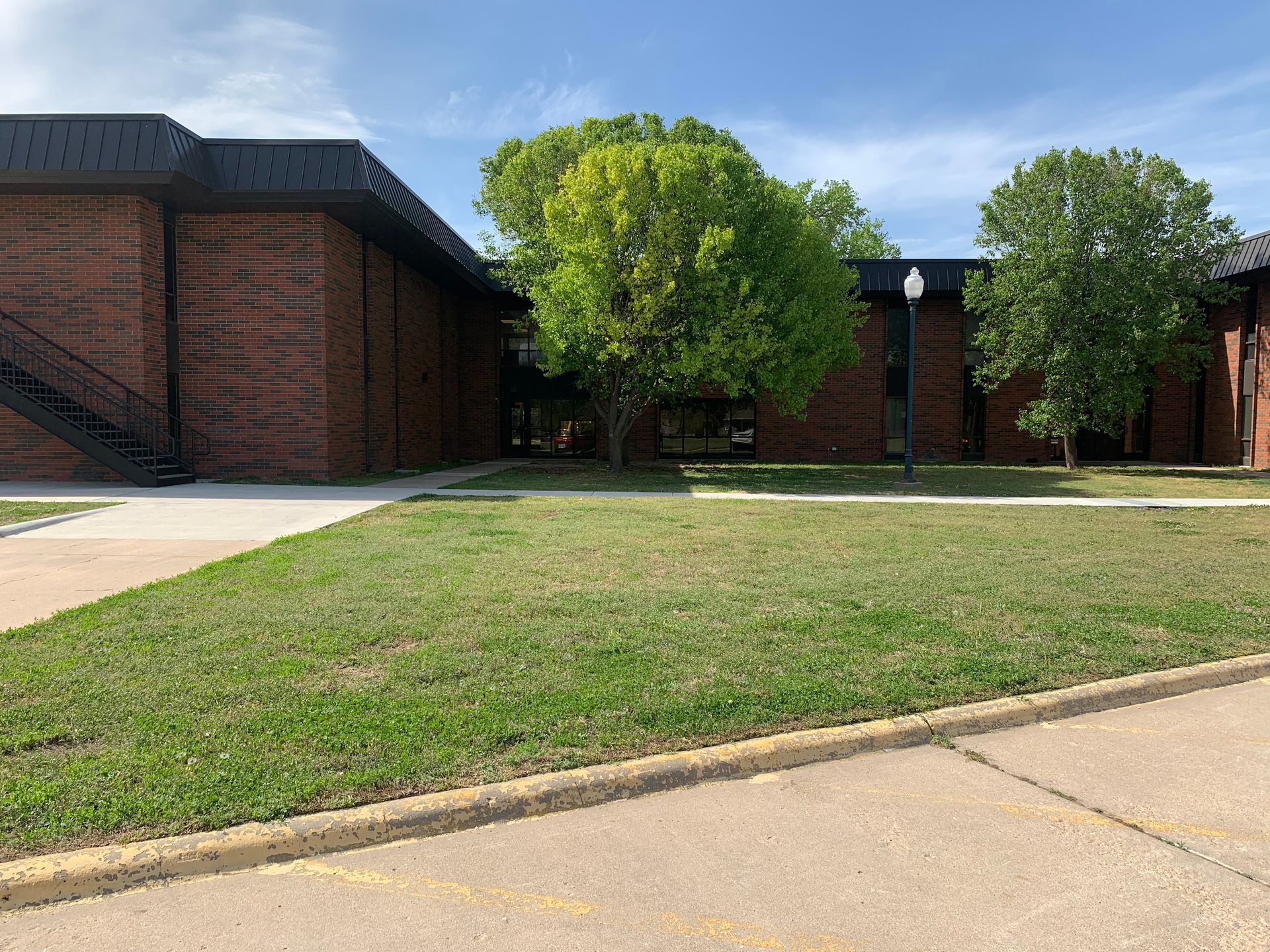 Brick buildings with a black roof and trees on a grassy lawn with a sidewalk.