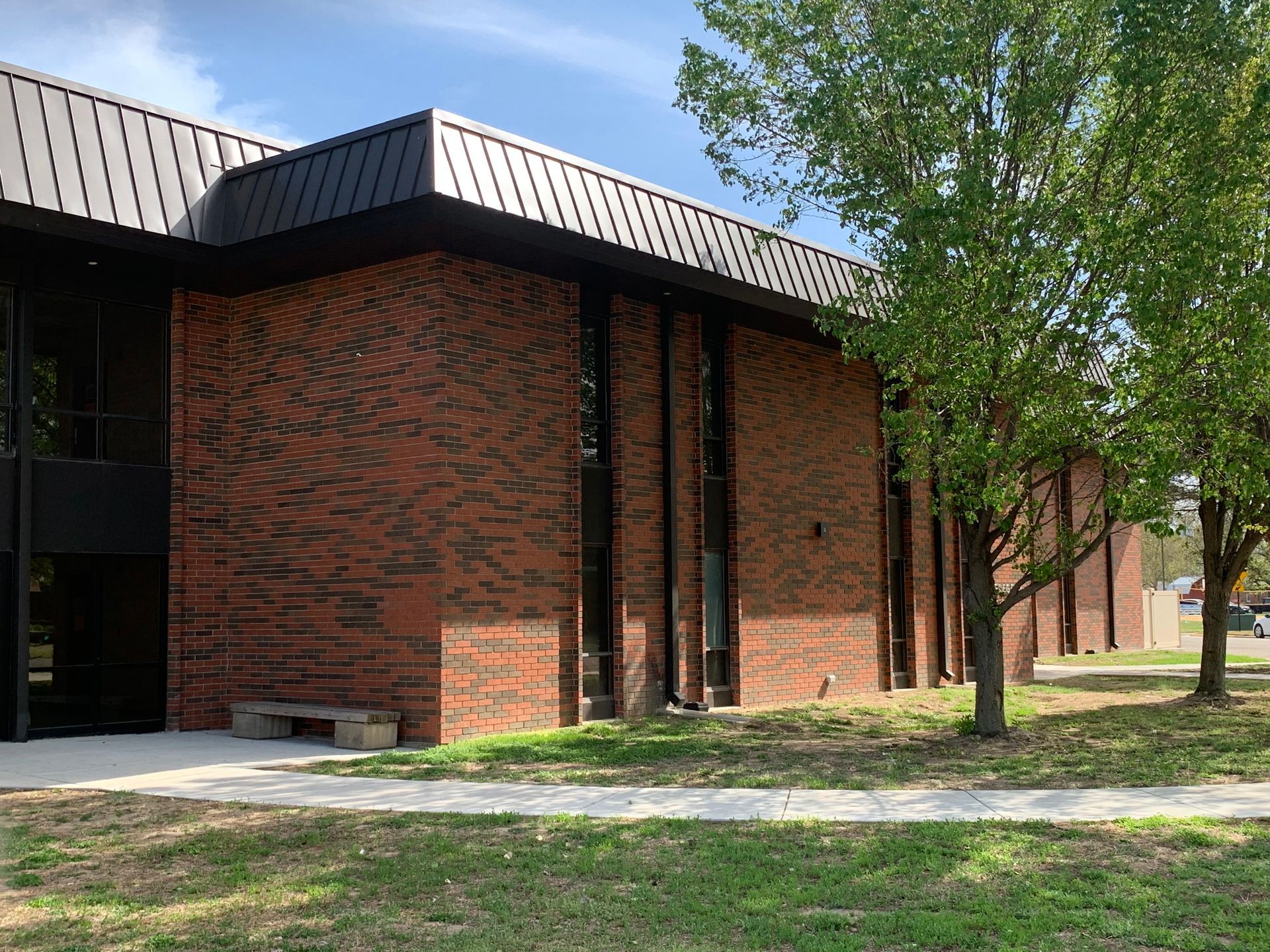 Brick building with black trim and tall windows, next to a grassy area with trees and a sidewalk.