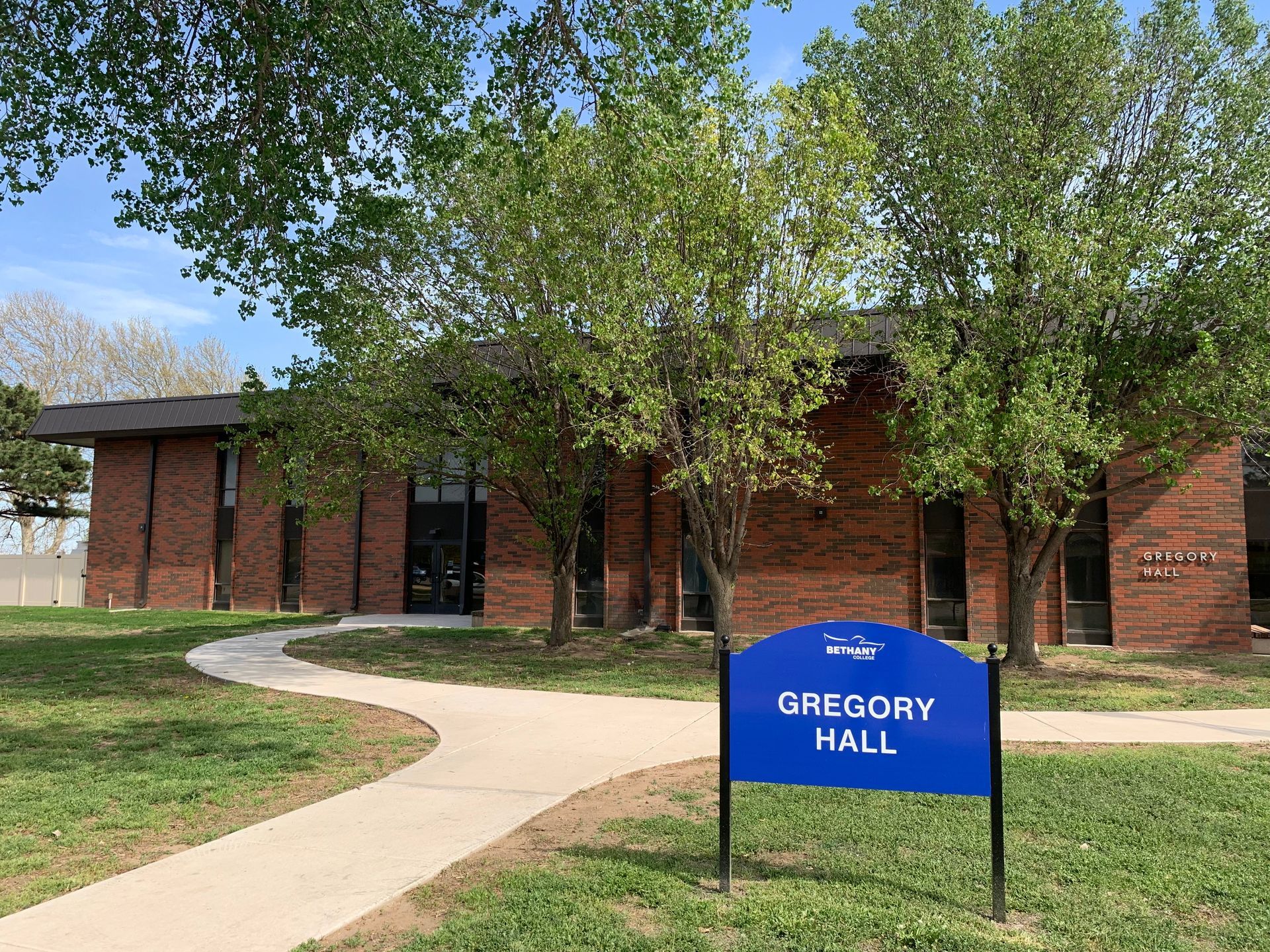 Gregory Hall building with blue sign, brick exterior, trees, and walkway on a sunny day.
