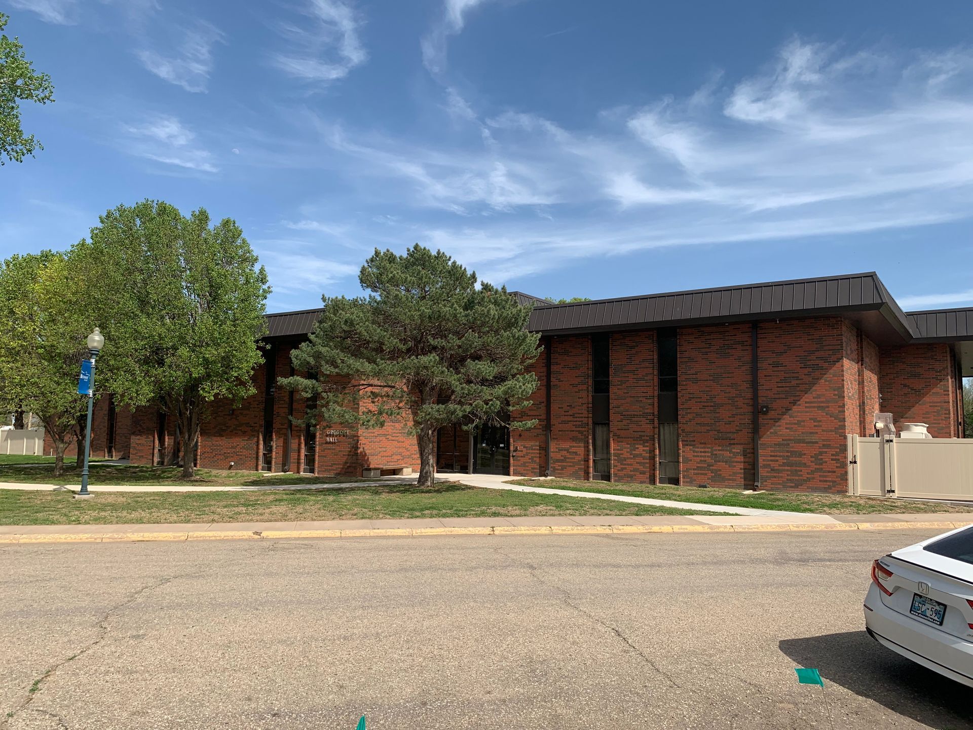 Brick building with dark roof under a partly cloudy sky. Trees and a street in front.