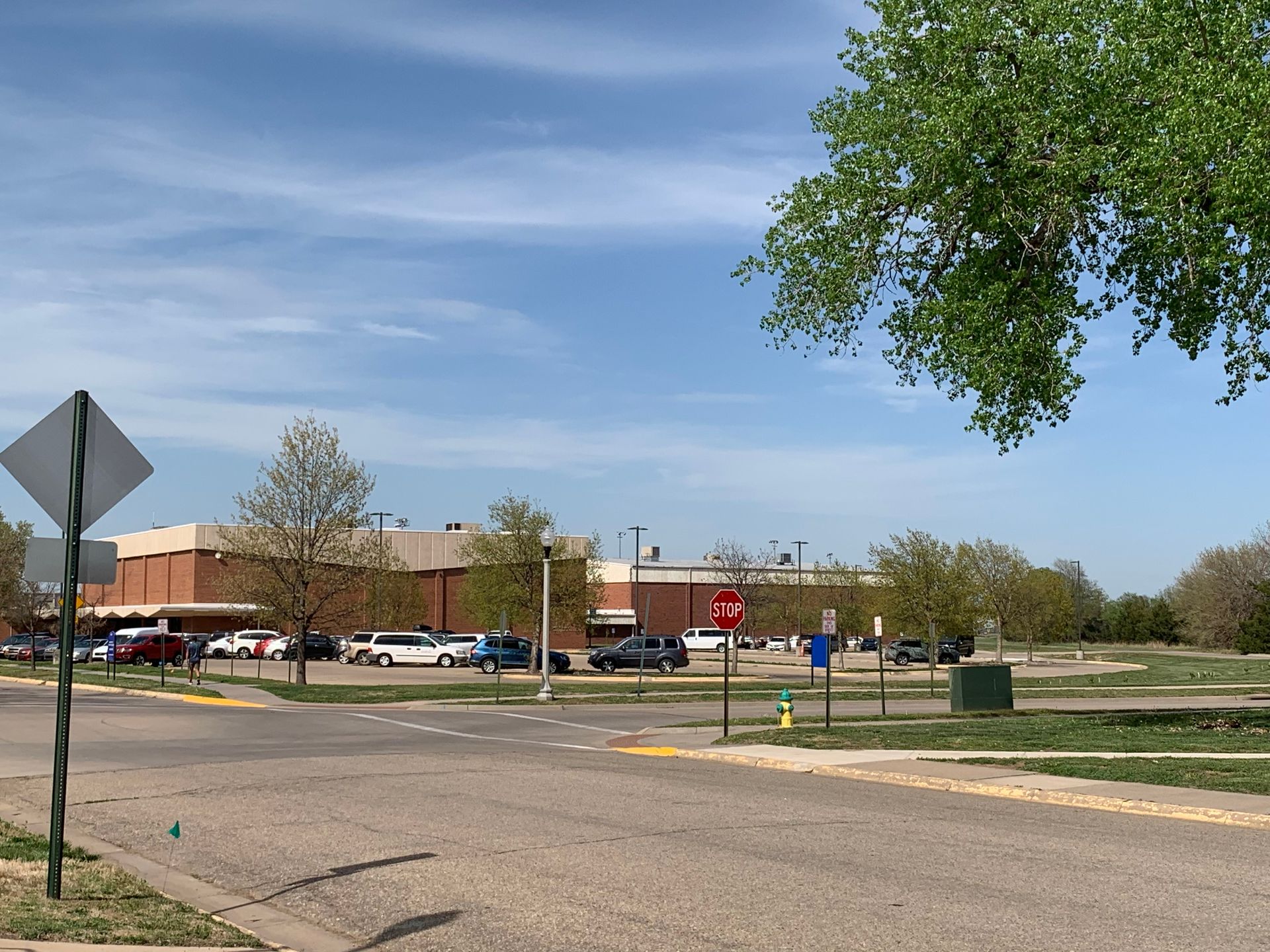 Parking lot with vehicles and building under a blue sky, road signs in foreground, tree on right.