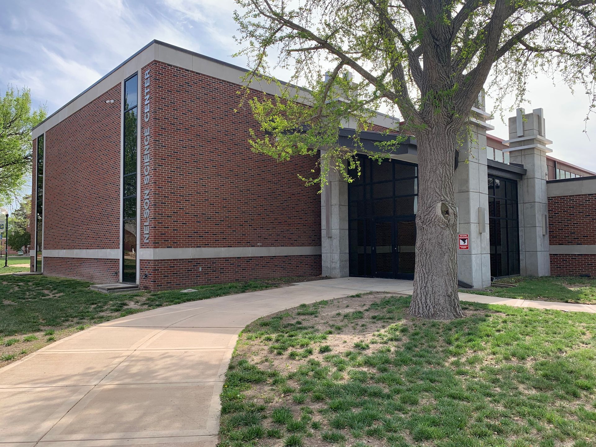 Brick building with a concrete walkway, tree, and black entrance; sunny day.