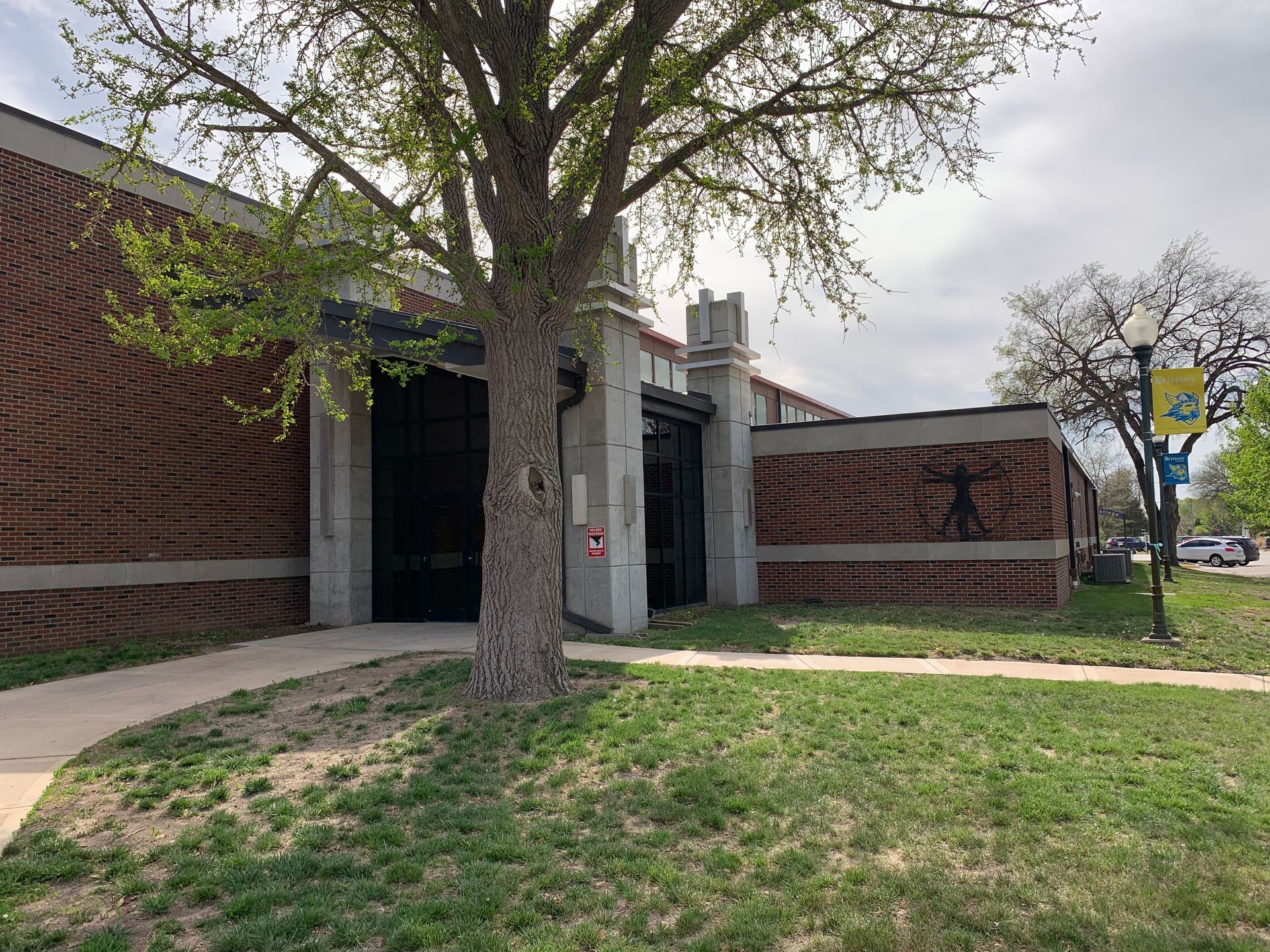 Brick building with arched entrance, tree in front, sidewalk, grassy lawn, cloudy sky.