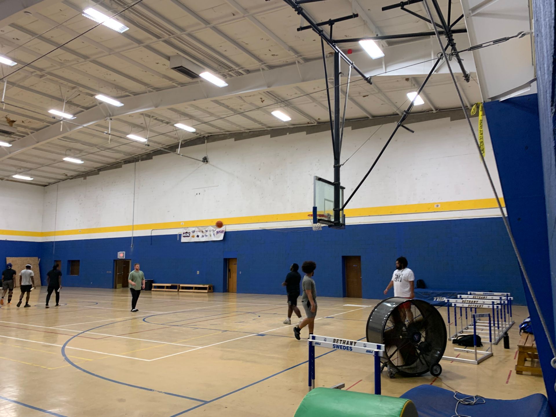 Basketball game in a gymnasium. People are playing; ball near hoop. Blue walls, yellow stripe, and hurdles visible.