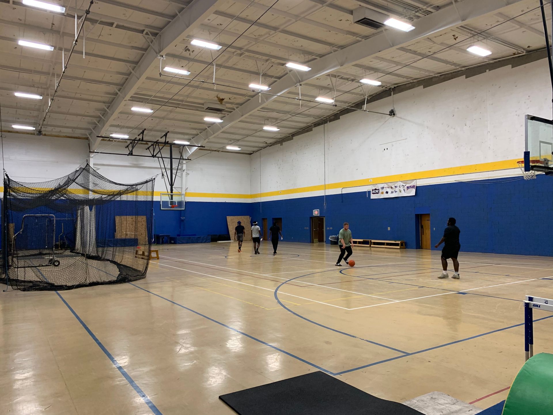 Basketball court with players. Blue and yellow walls, a net structure, and players dribbling and walking.