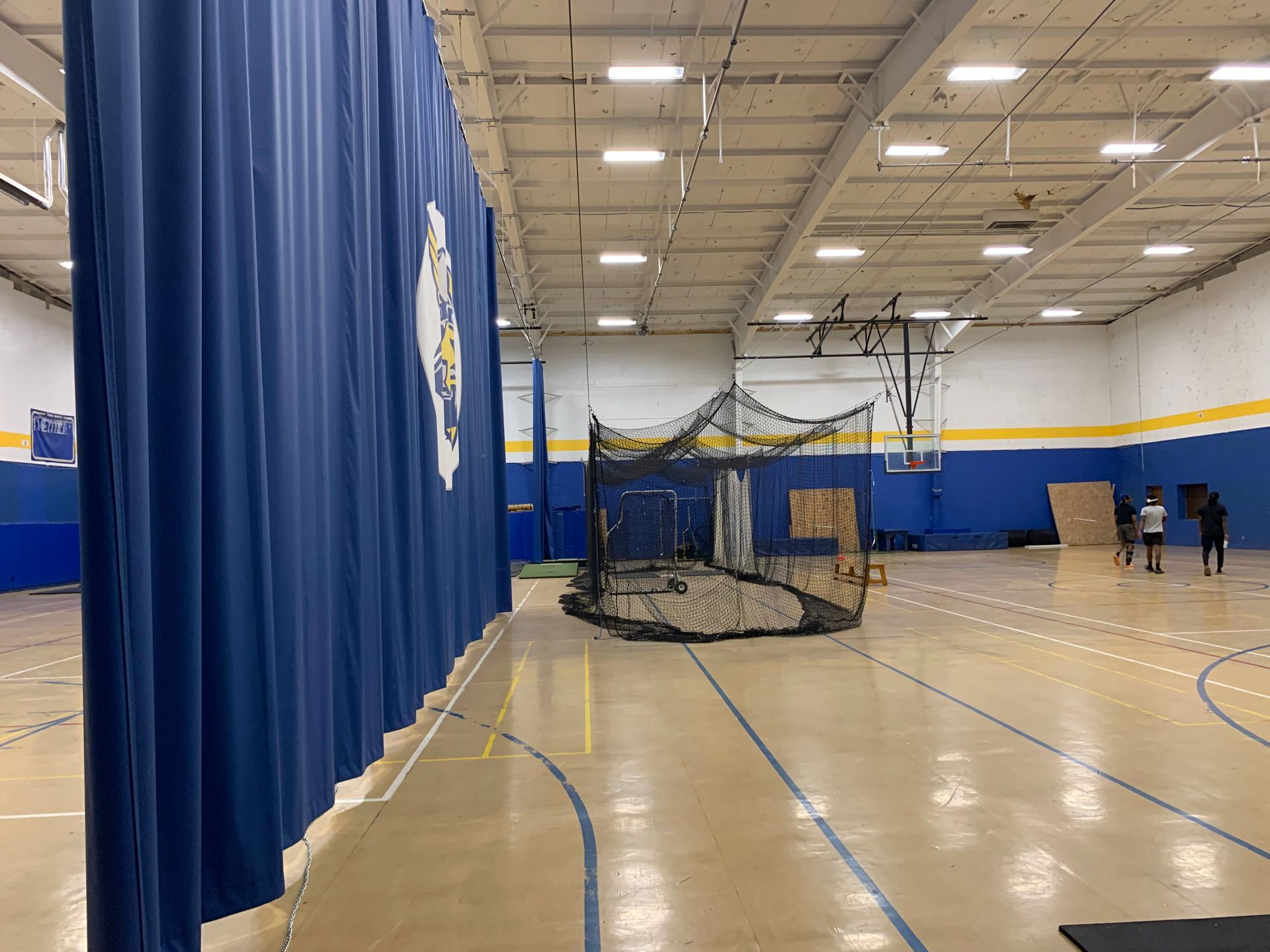 Indoor gymnasium with a blue curtain, batting cage, and two people walking.