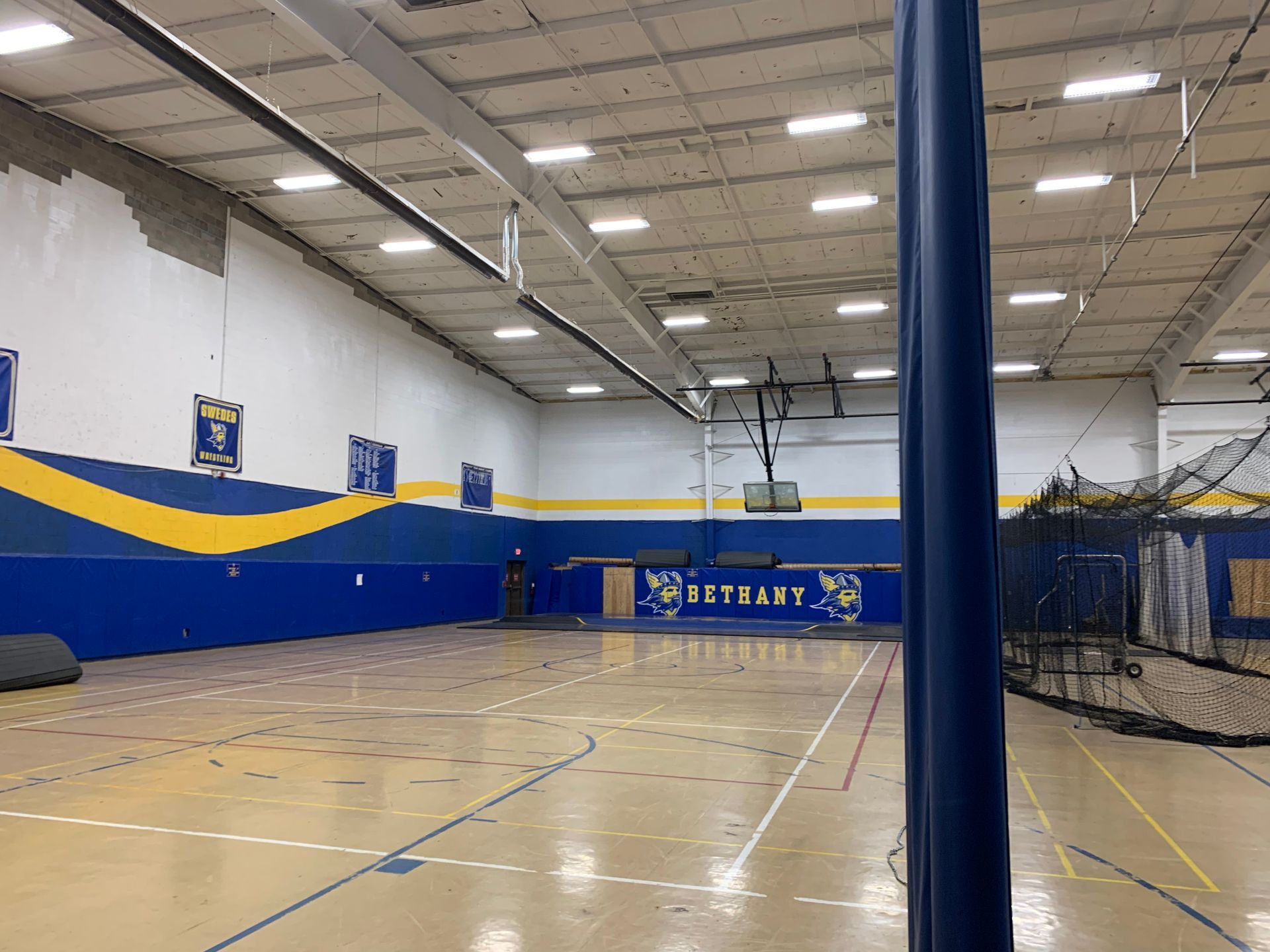An empty gymnasium with blue and yellow accents. A basketball hoop and a batting cage net are visible.