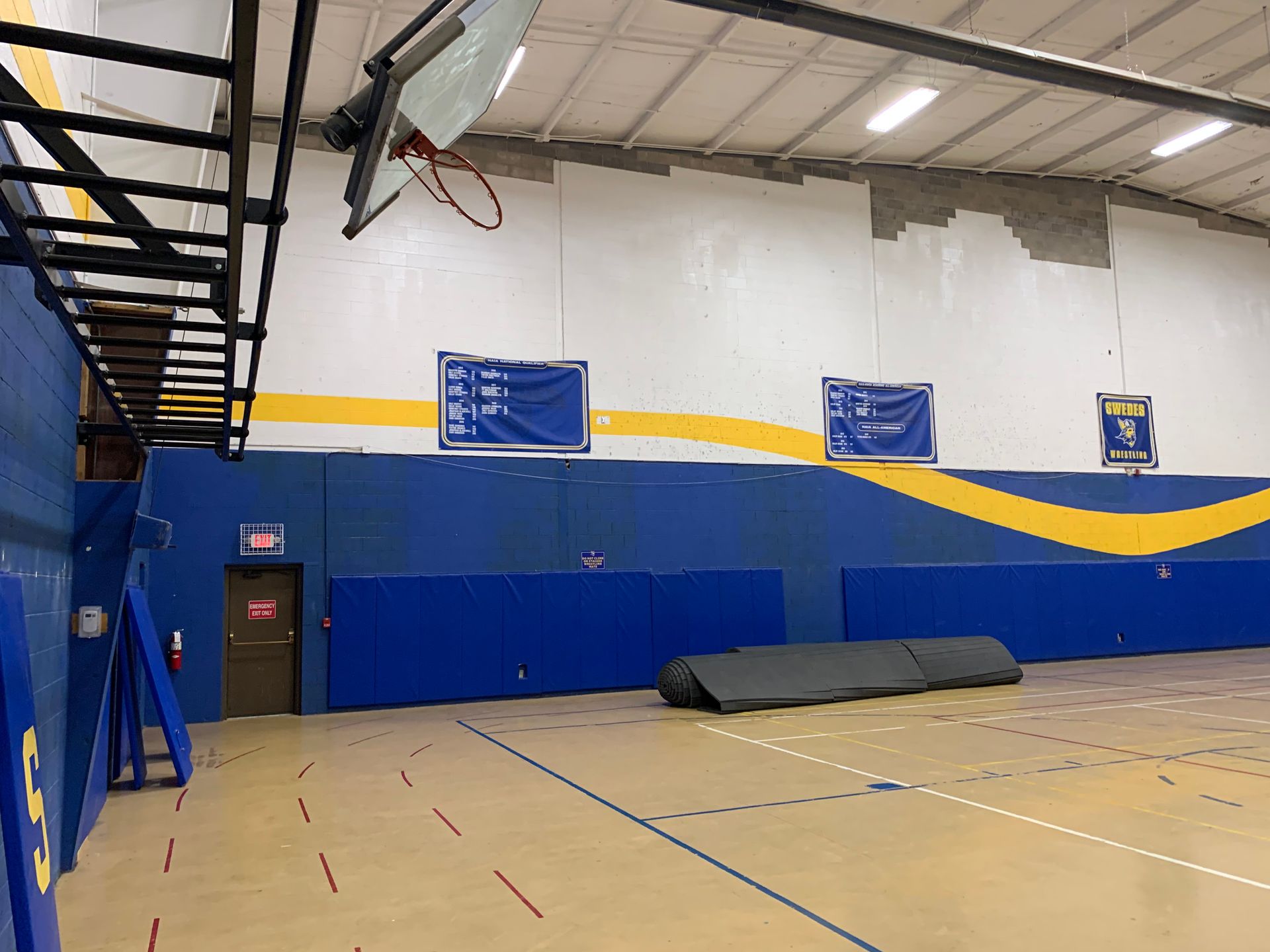Gymnasium interior with blue and yellow wall padding, basketball hoop, and climbing structure.