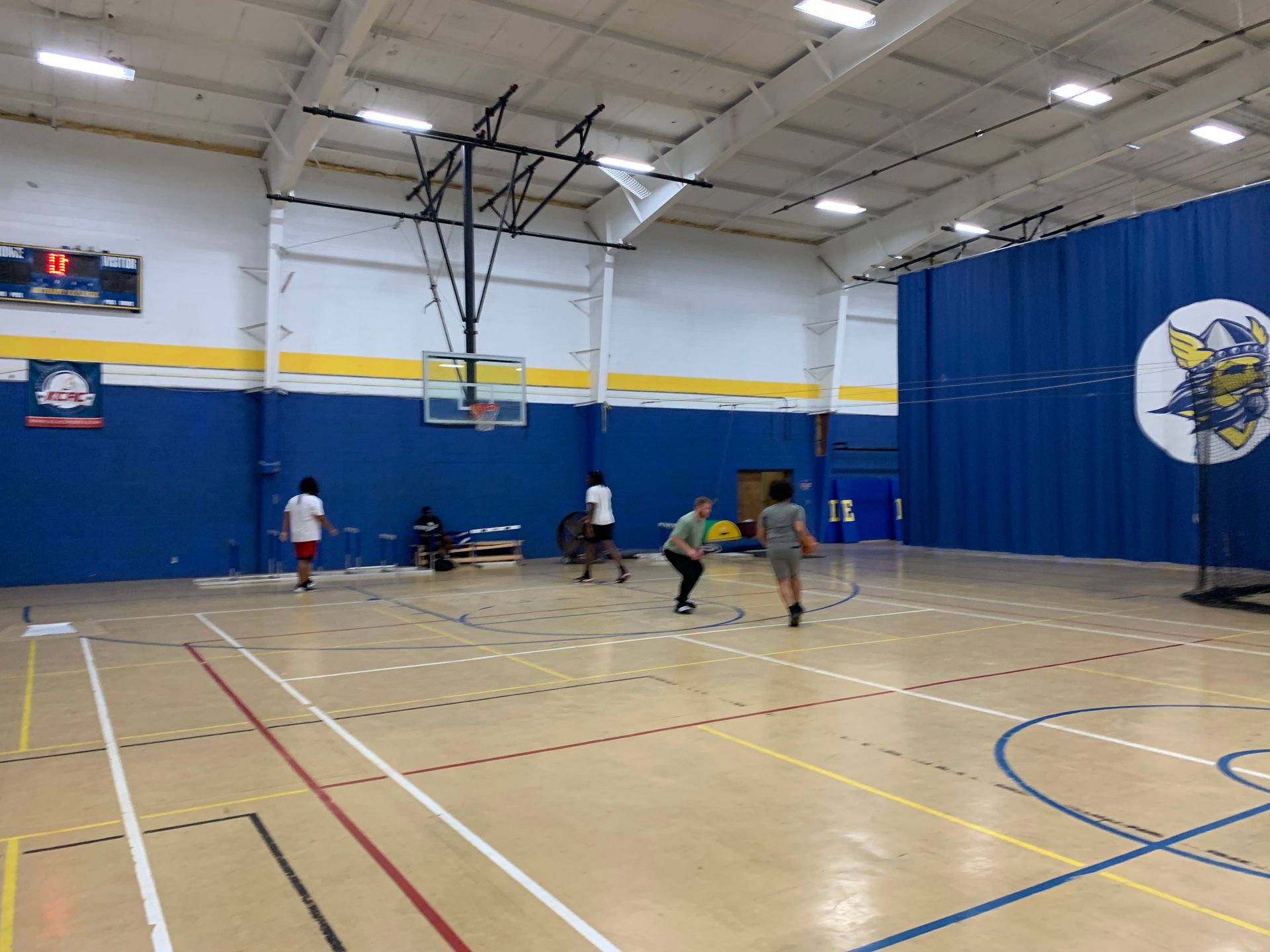 People playing in a gymnasium with basketball hoop. Blue and yellow walls.