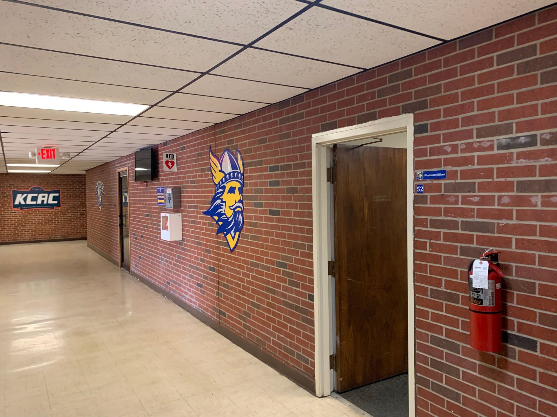 Hallway with brick walls, doors, school logo, and fire extinguisher.