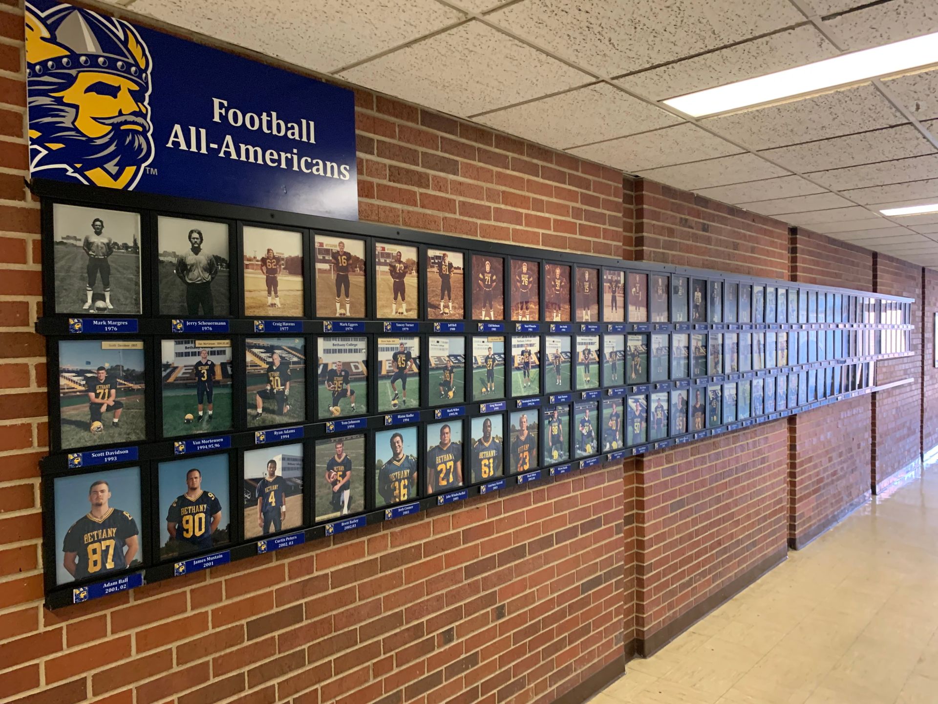 Hallway wall displays framed photos of football All-Americans against a brick wall, with a team logo.