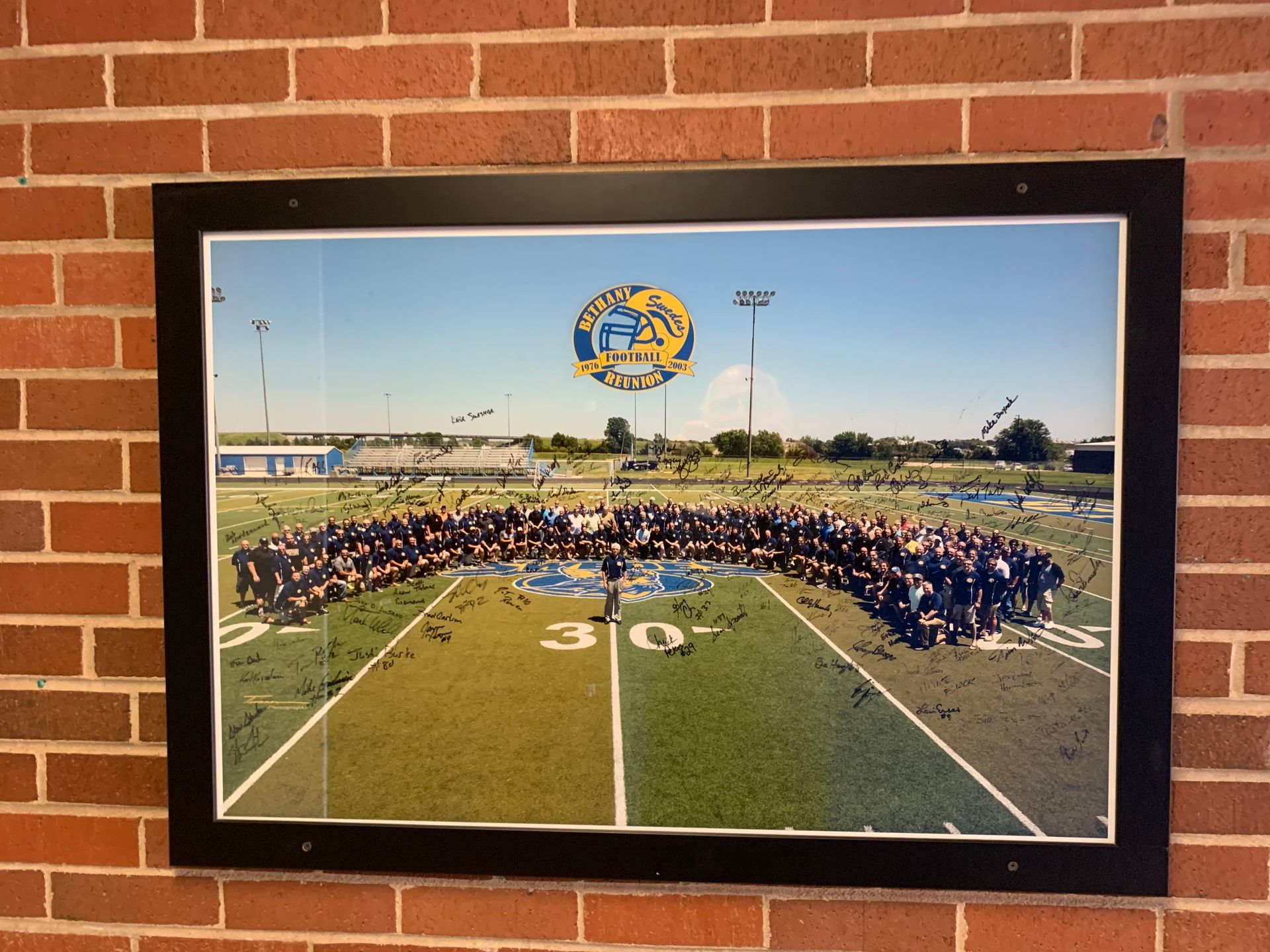 Framed group photo on brick wall. People on football field beneath logo. Sky and stadium in background.