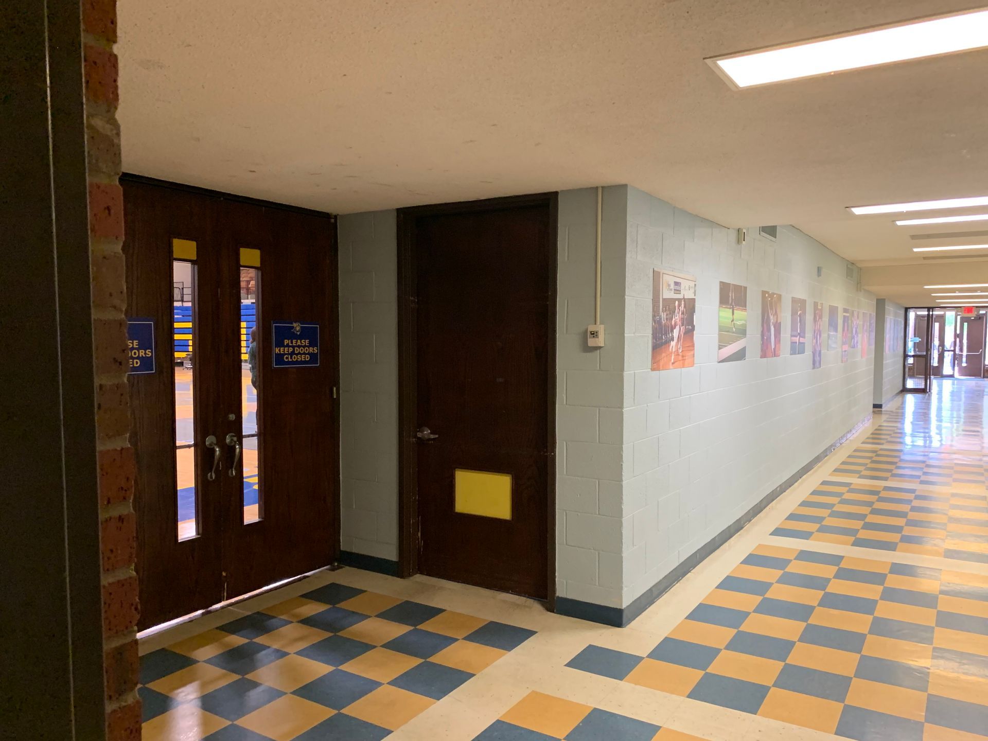 Hallway with doors on the left, blue and yellow checkered floor, posters on the right, and bright overhead lights.