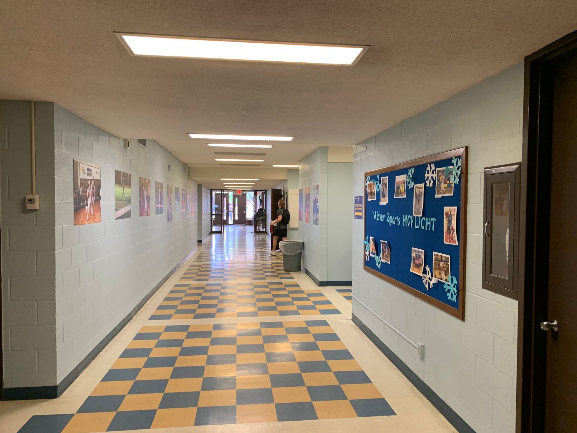 School hallway with blue and yellow checkered floor, light blue walls, posters, bulletin board, and doors.
