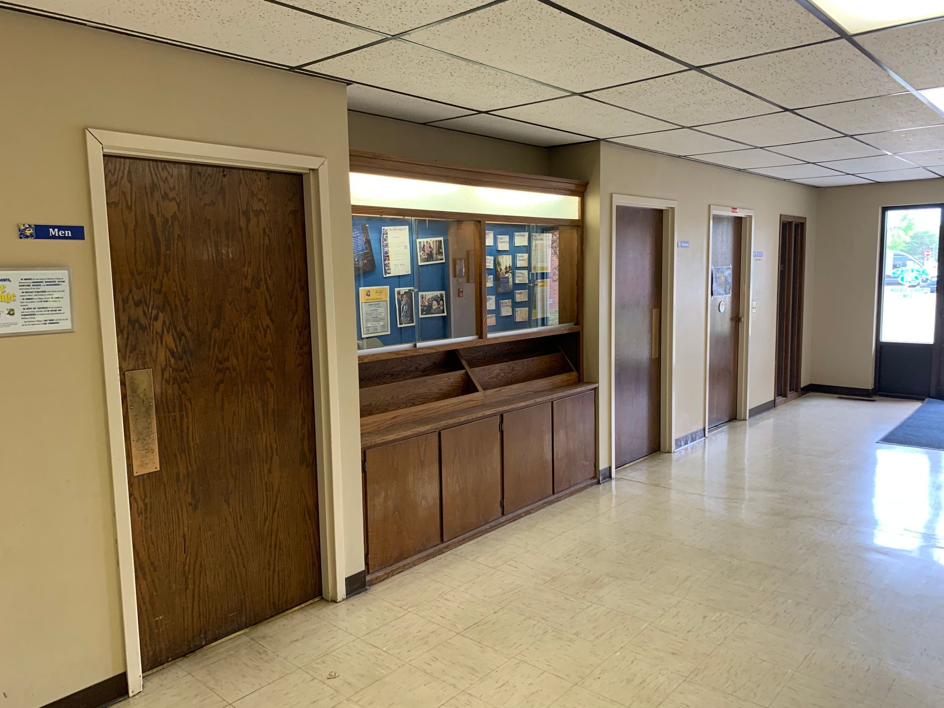 Hallway with doors, bulletin board, and ceiling tiles. Light brown walls and wooden doors.