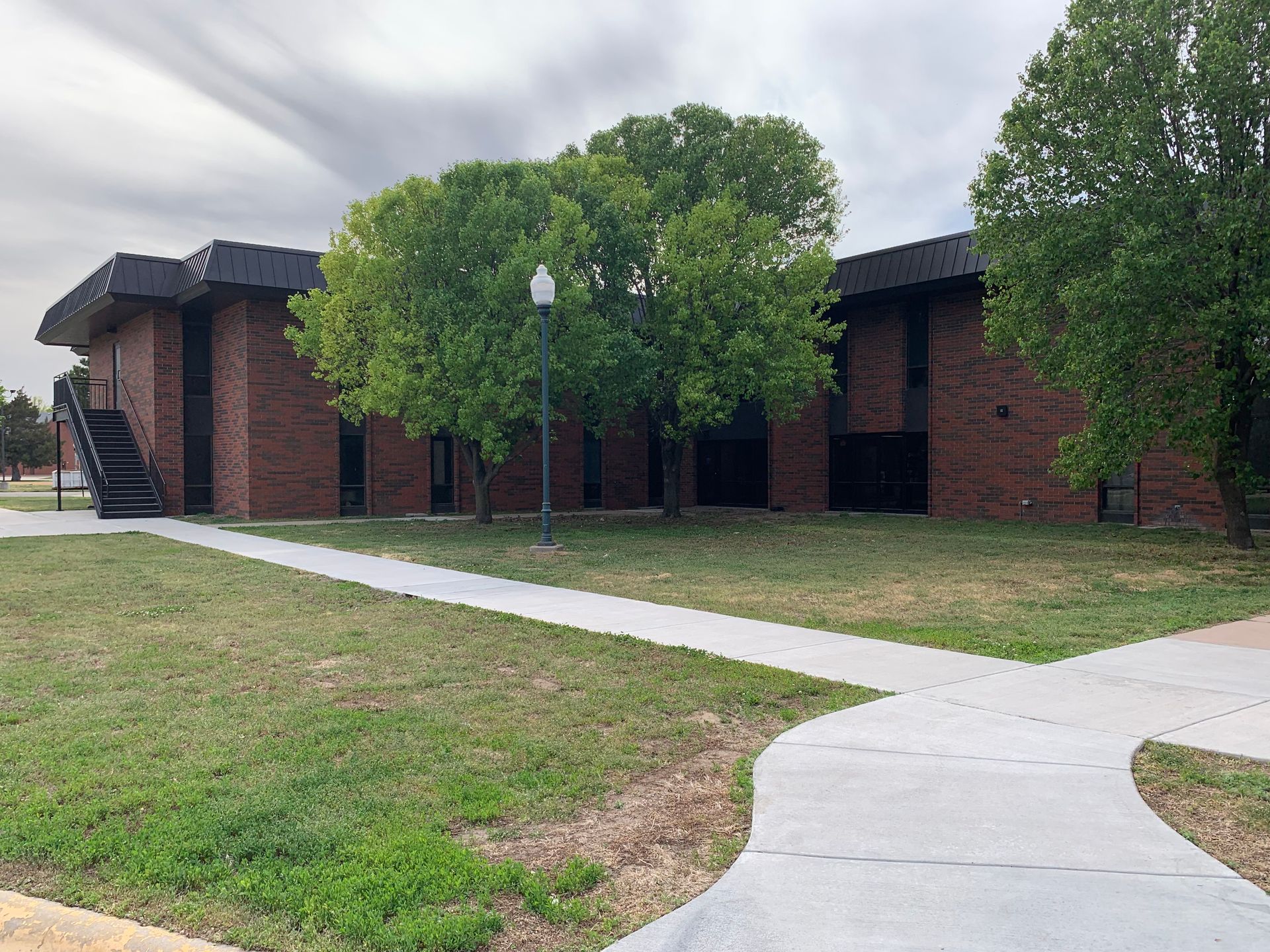 Brick building with a dark roof and trees, a sidewalk leading up to the entrance under a cloudy sky.
