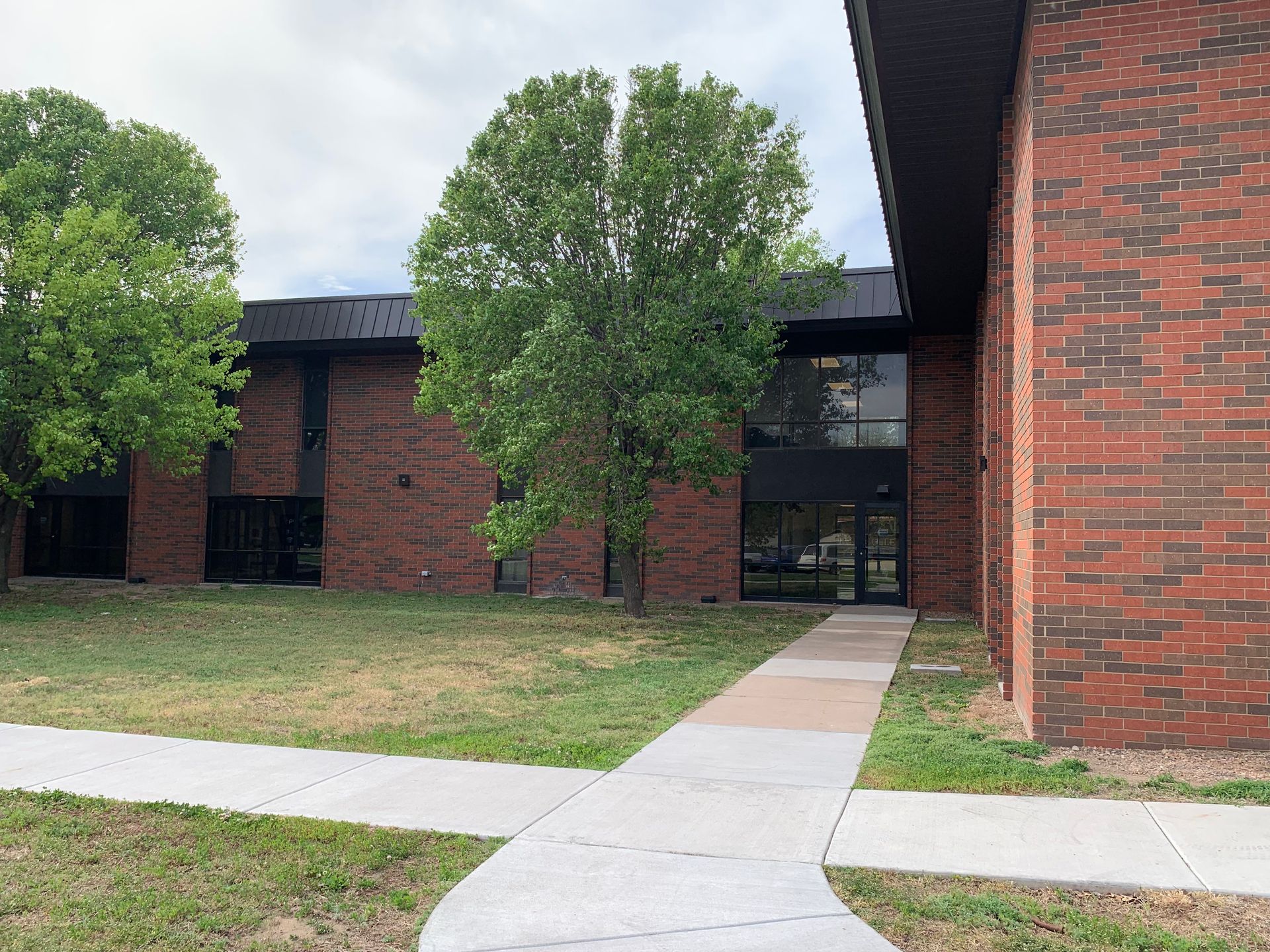 Red brick building with a concrete walkway leading to the entrance, a tree in the center, and a cloudy sky.