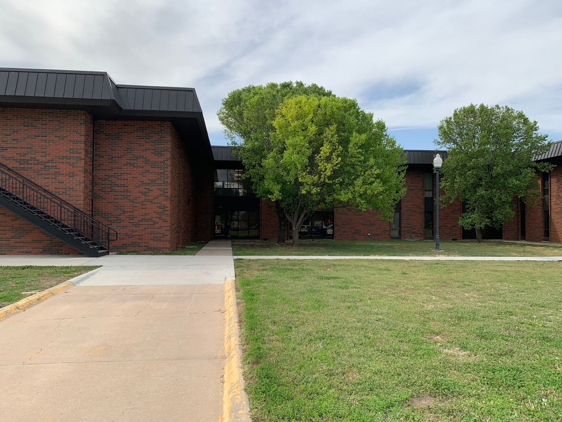 Brick building with a dark roof and trees, a sidewalk leading to the entrance, under a cloudy sky.