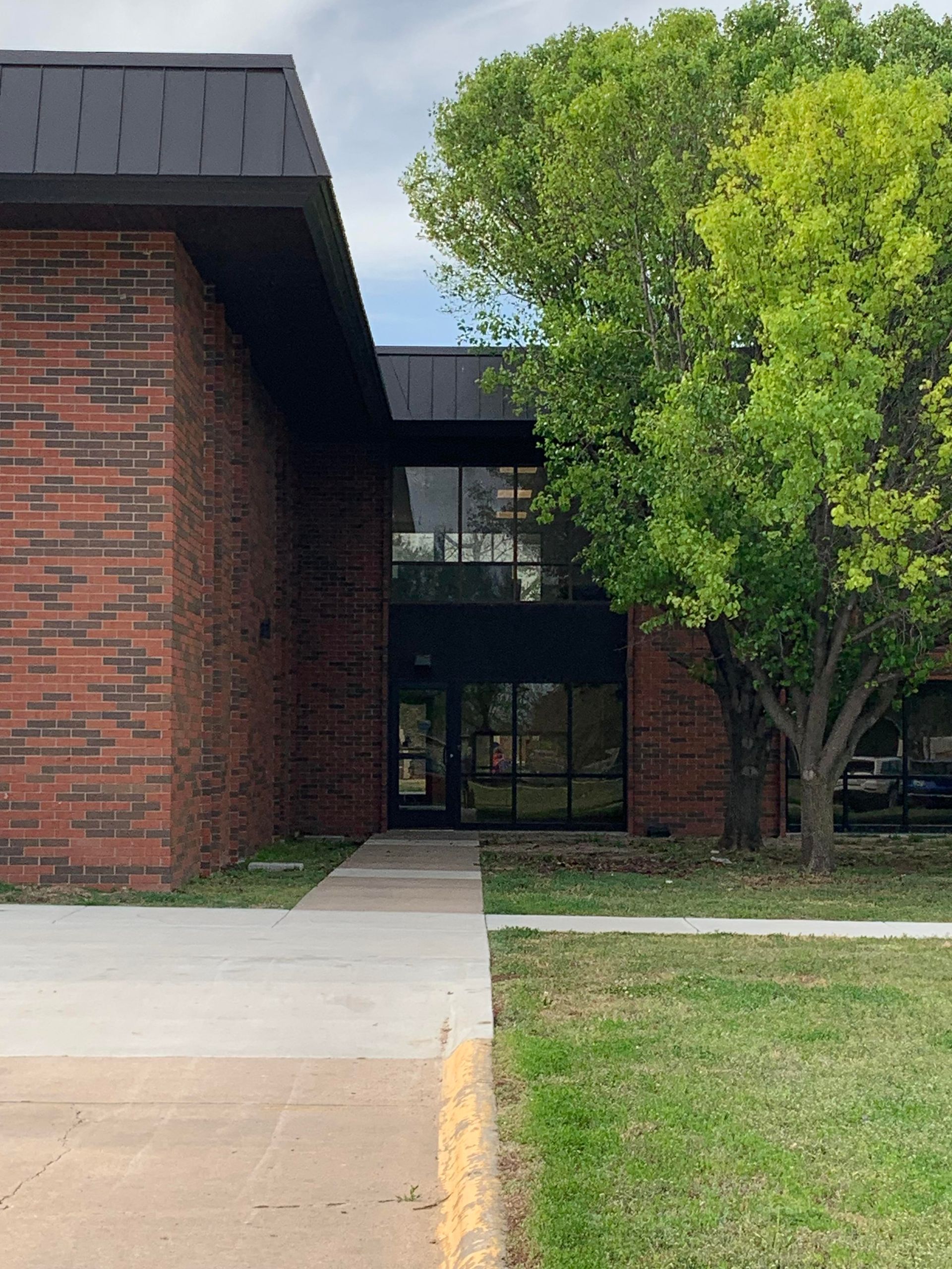 Brick building with black trim and glass entryway, sidewalk, and green grass with tree.