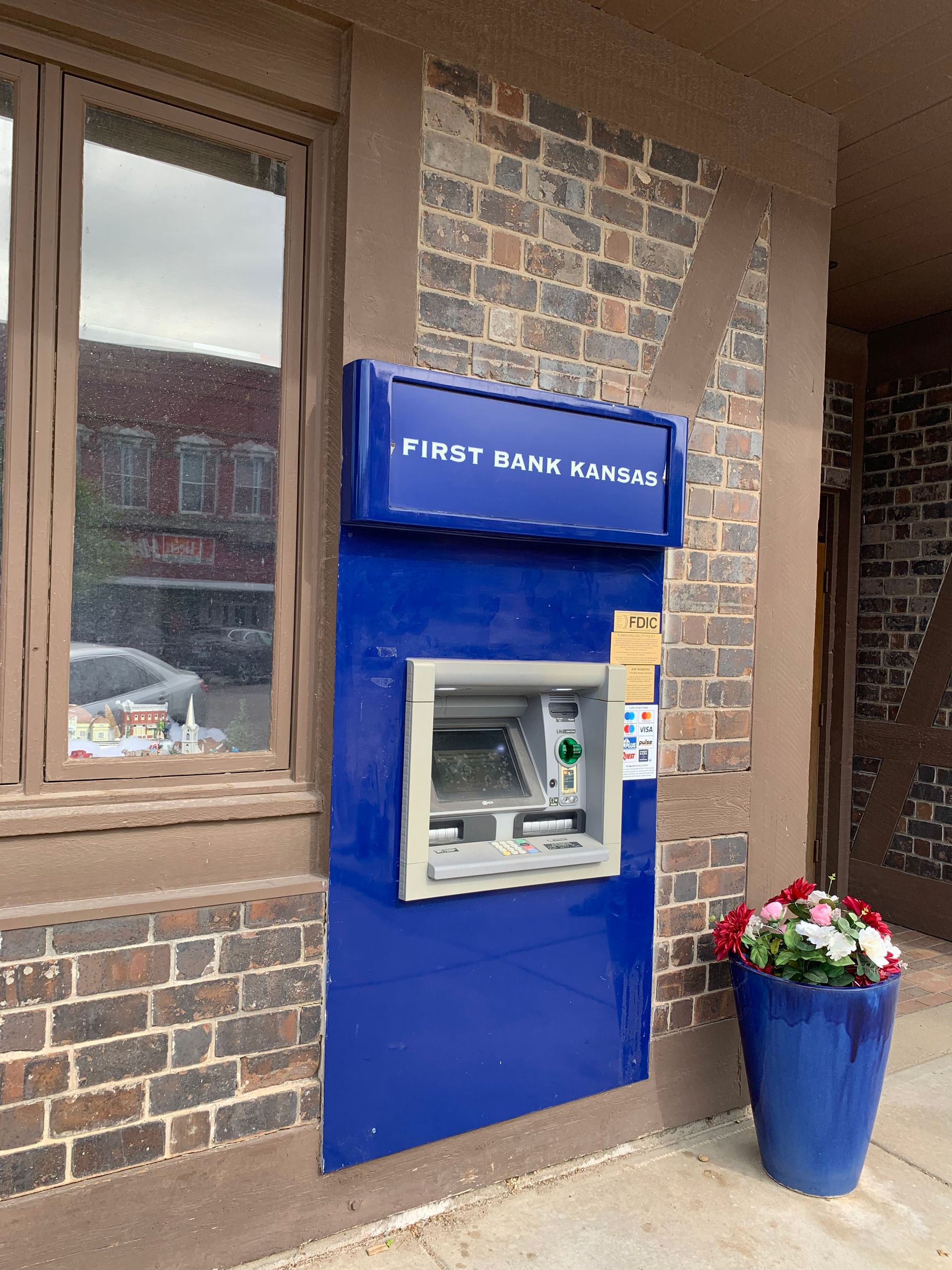 Blue ATM for First Bank Kansas on a brick building, with a flower pot.