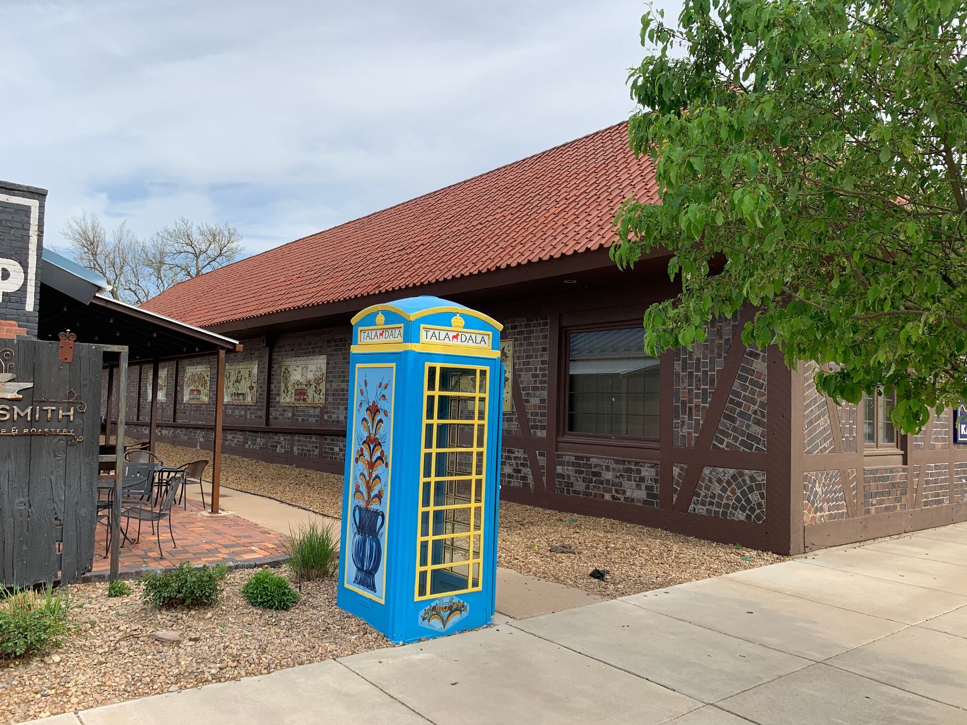 Blue painted phone booth stands on a gravel path, in front of a brown building with a red roof.