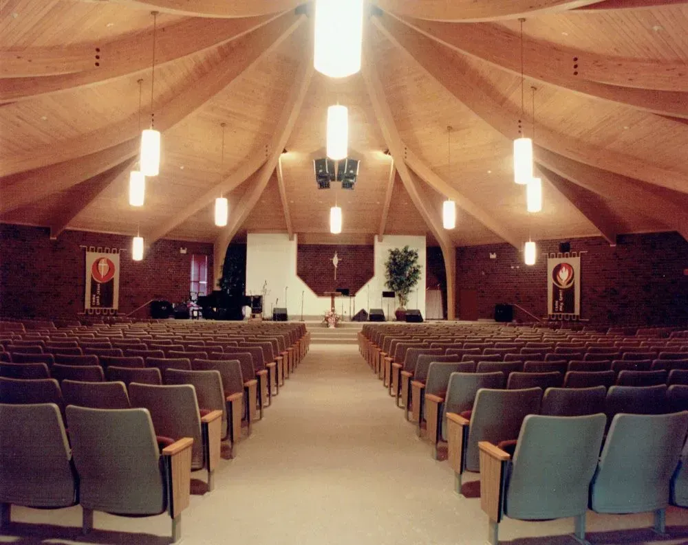 Interior view of a church auditorium with rows of seating facing a stage; wooden ceiling, brick walls.