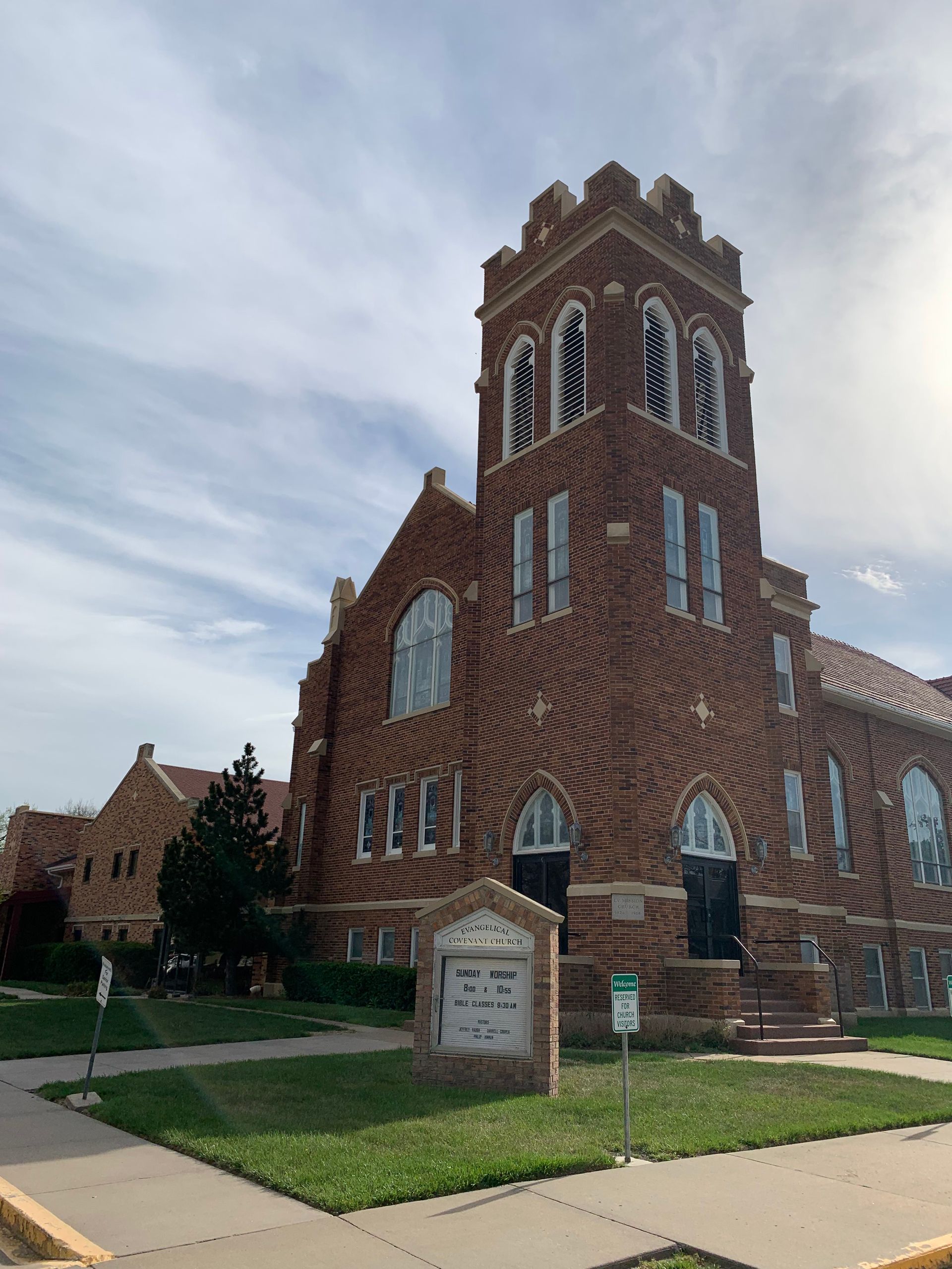 Brick church with a tall tower, arched windows, and a sign in a grassy setting under a cloudy sky.