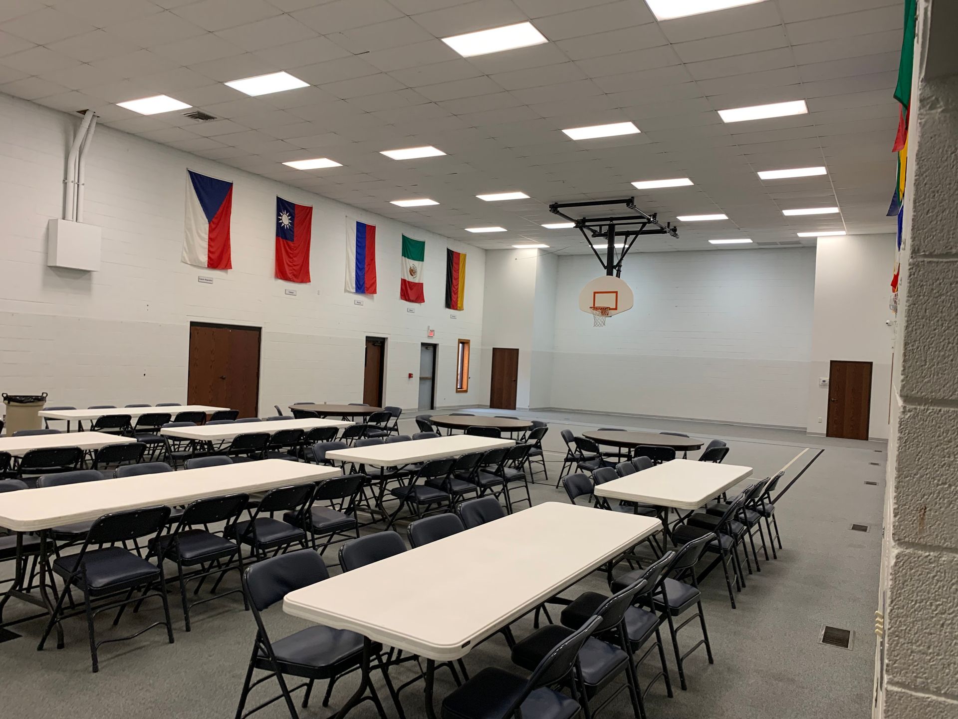 Large room with tables, chairs, and basketball hoop. Flags hang on the wall.