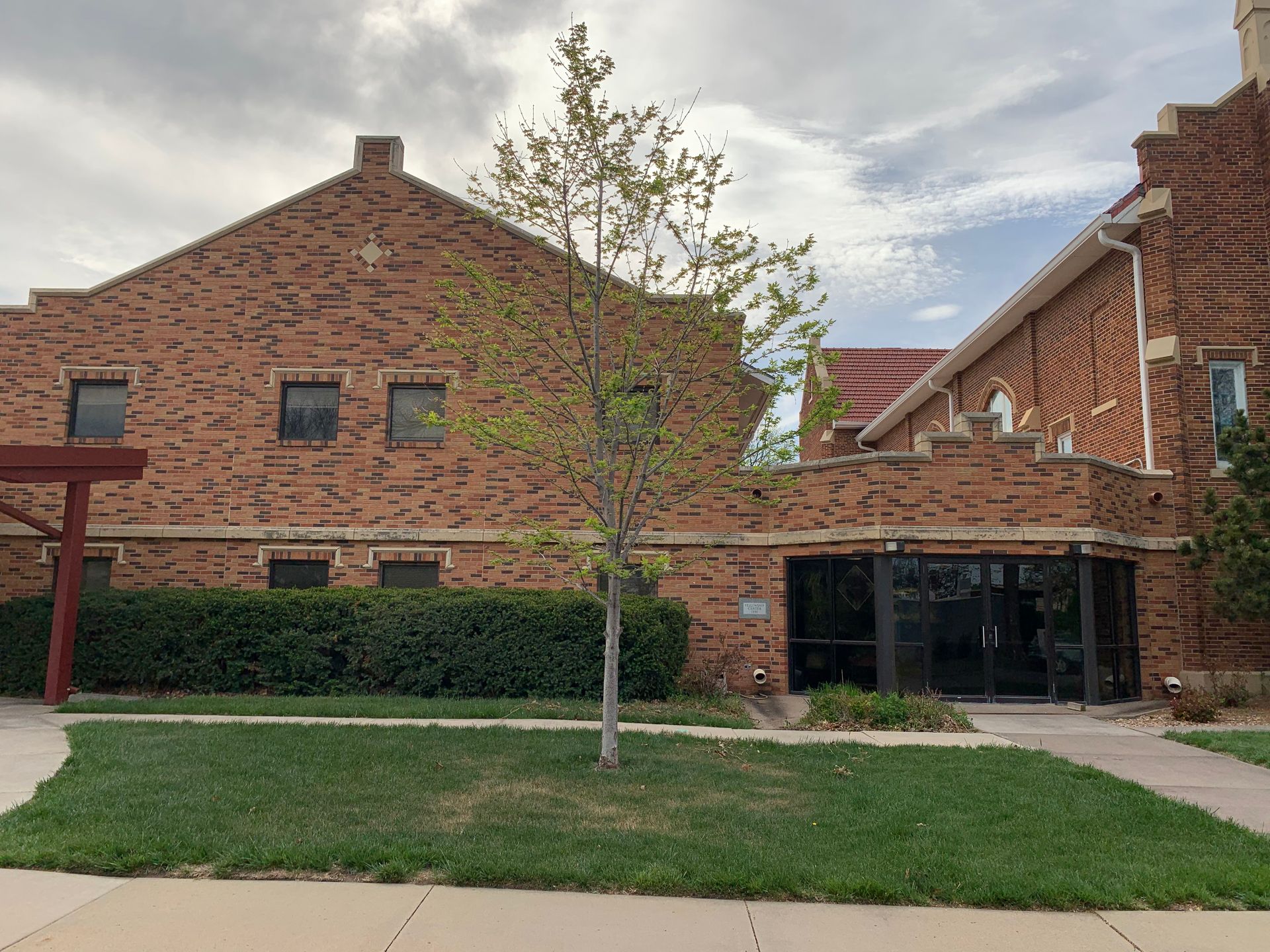 Brick church building with a tree in front and green grass. Cloudy sky.