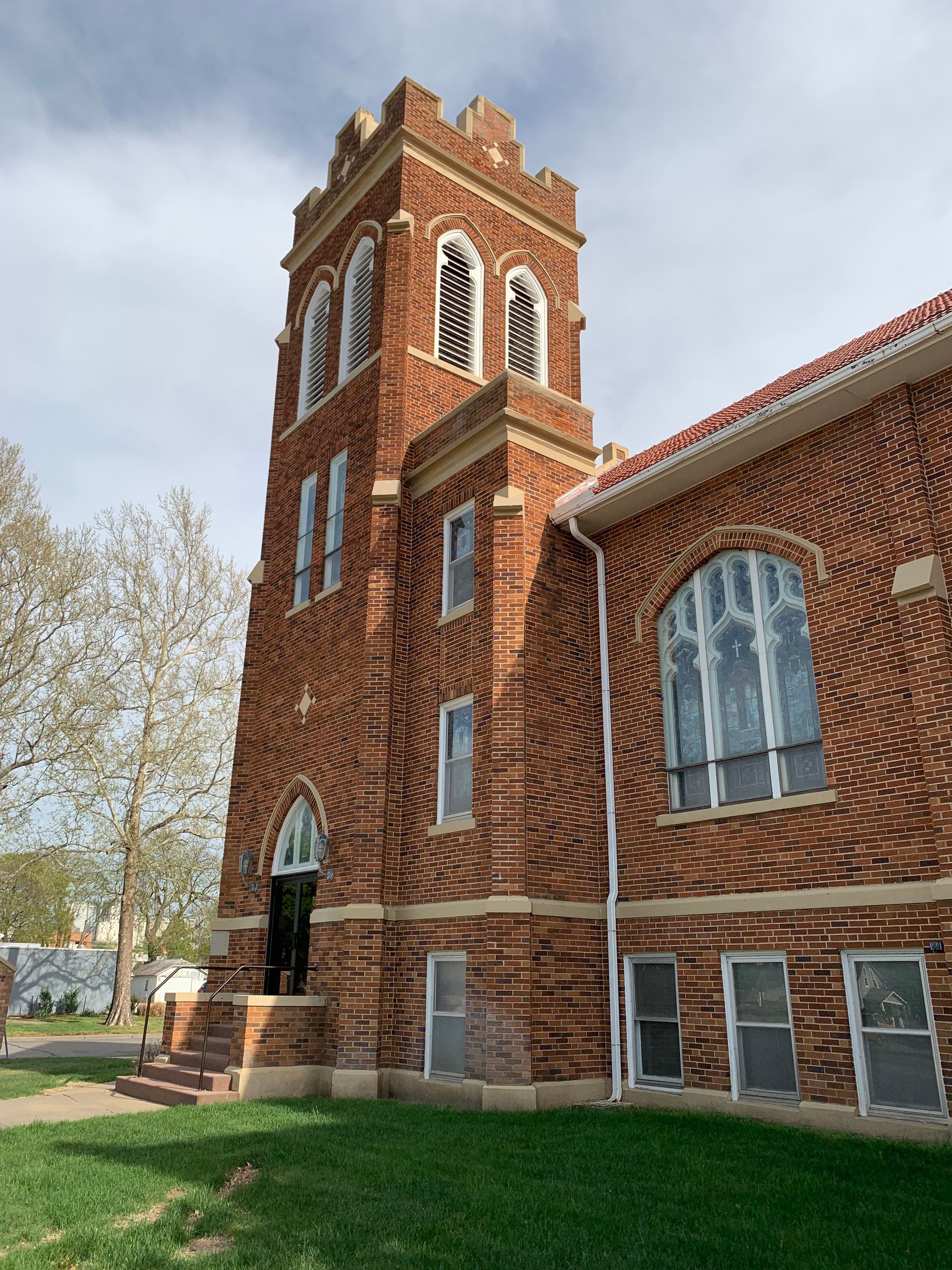 Red brick church tower and building, arched windows, green lawn, cloudy sky.
