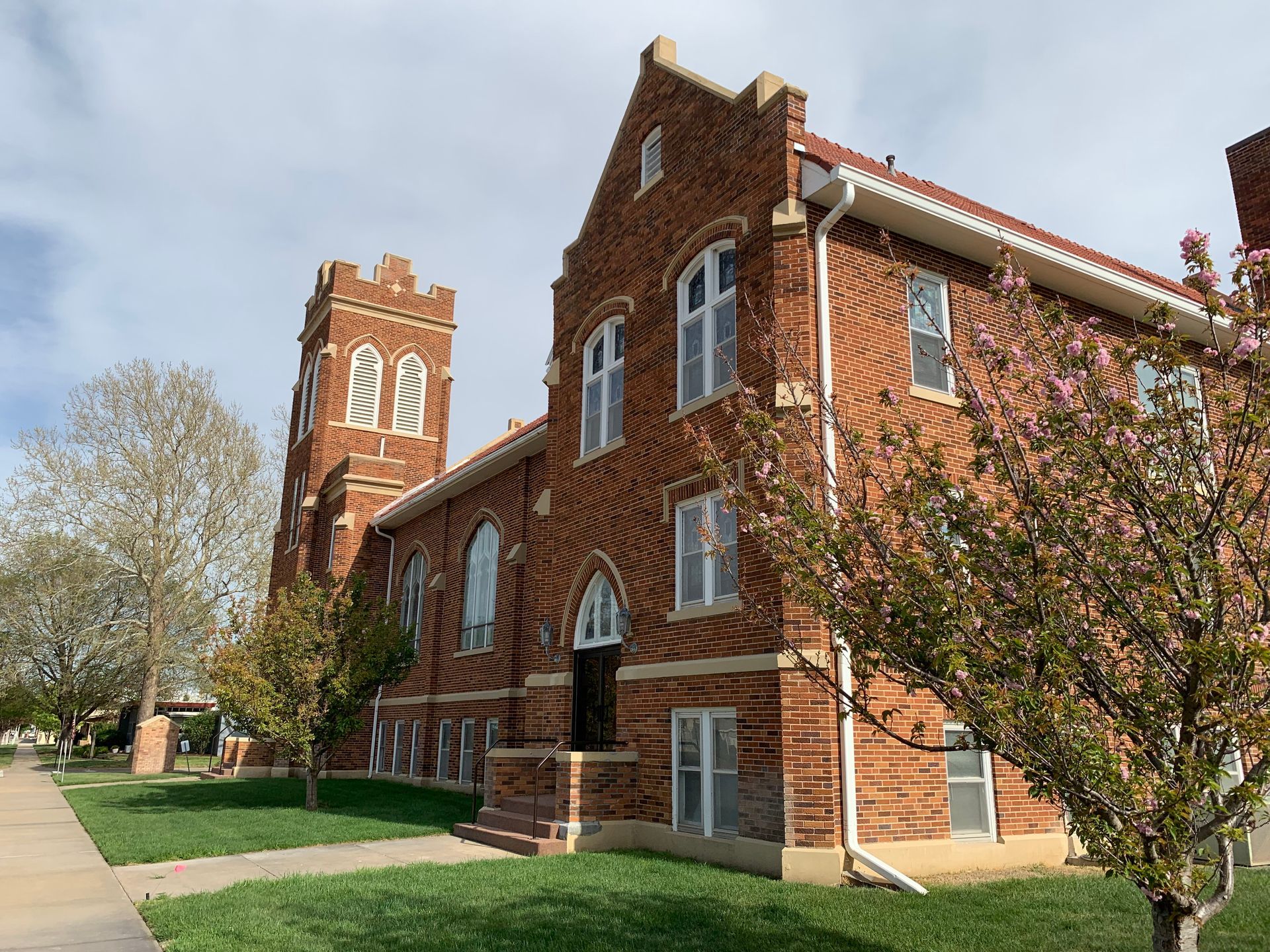 Brick church building with a tower on a sidewalk, under a partly cloudy sky.