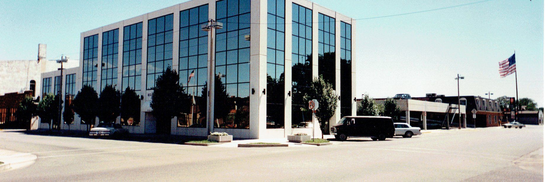 A glass-walled building with trees reflected in its windows, with an American flag waving in the distance.