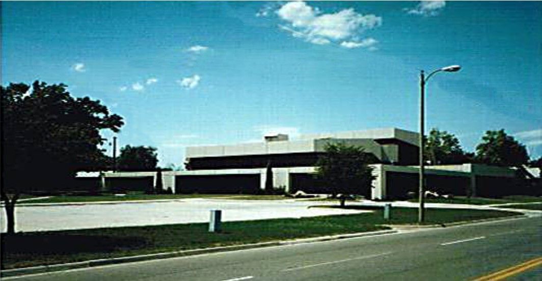 A low, modern building with a flat roof, large windows, and a parking lot on a sunny day.