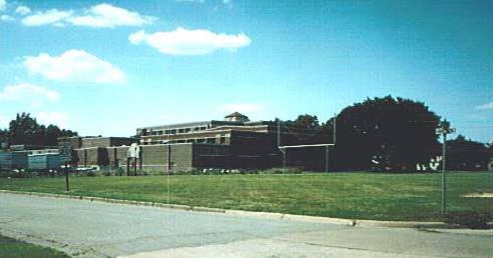 Building with multiple floors, brown brick exterior, on a grassy field under a blue sky with clouds.