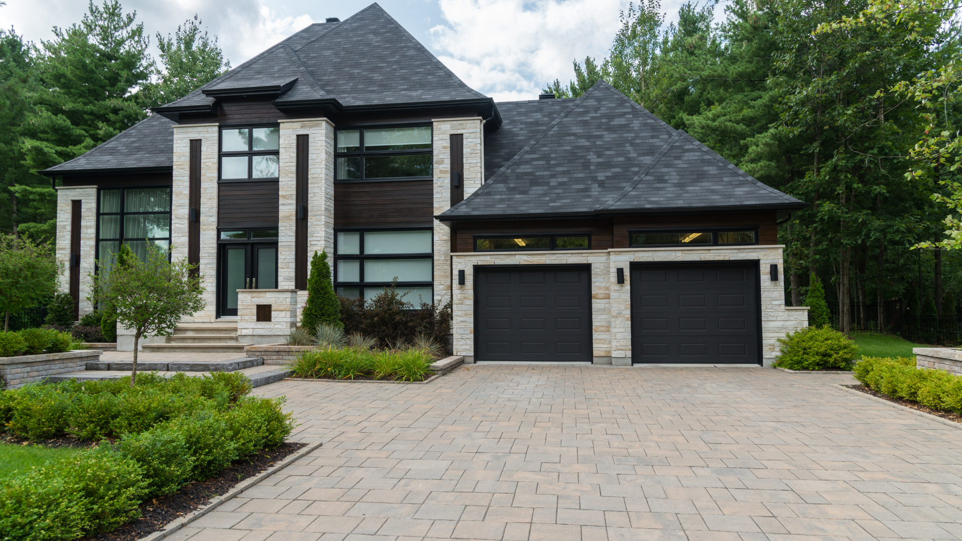 Modern house with black roof, gray stone accents, black garage doors, and brick driveway.