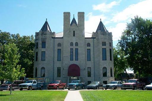 Stone building with arched windows, turrets, and a red awning. Cars parked on grass in front.