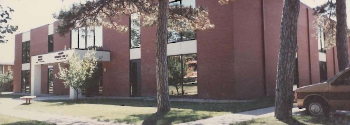 A two-story brick building with large windows. Trees and a car are in the foreground.