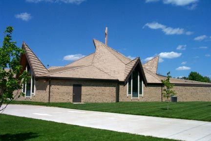 Tan brick church with distinctive roof peaks, cross, and a pathway on a sunny day.