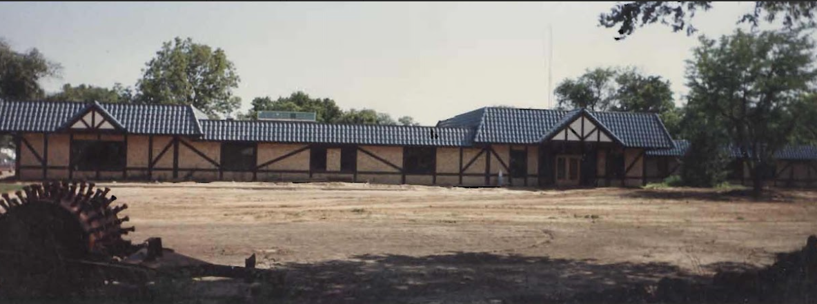 A long, single-story building with a dark roof and light-colored walls, surrounded by trees and a brown field.