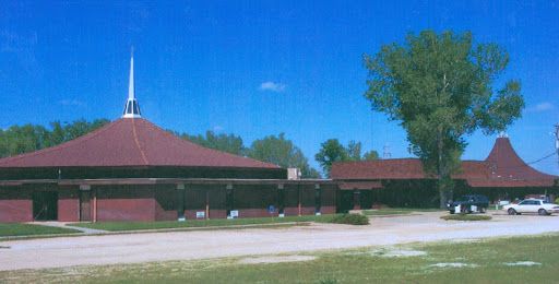 Brown brick church with a tall steeple, set in a grassy area with trees and blue sky.