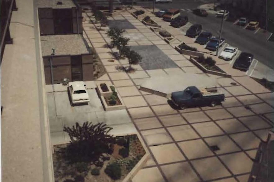 Overhead view of a paved area with parked cars and trees. Buildings flank the area, with a street in the background.