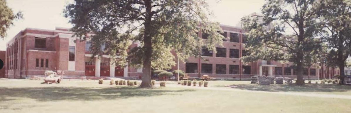 A brick school building with many windows and trees on a grassy lawn.