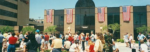 People gathered outside a brick building with American flags; the sky is blue.