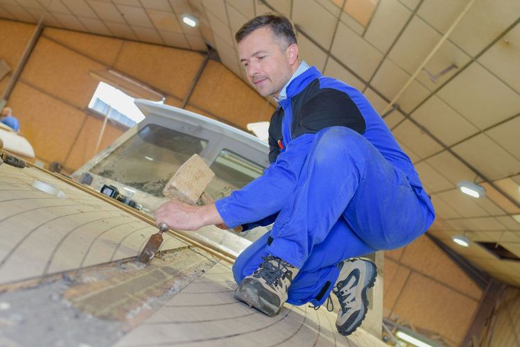 Man crouching down to scrape off old paint from the bow of a small boat so he can start the refinishing process